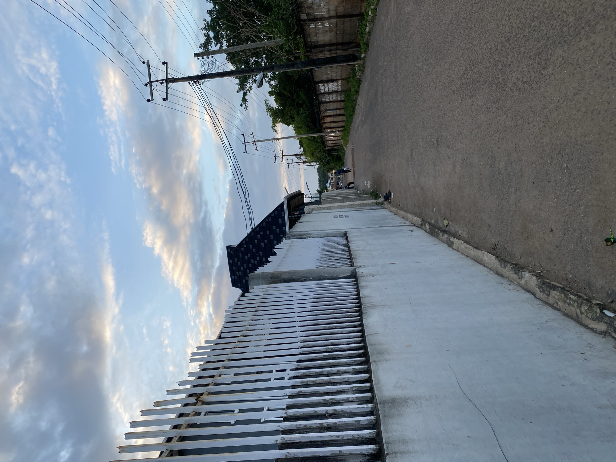 The image depicts a street scene in Abeokuta, Nigeria, under a cloudy sky. On the left, a white fence with vertical metal bars runs along a street. Behind the fence, a building with windows is visible, and a dark, angled roof with black shingles is also present. The fence appears somewhat aged and weathered.

The street itself is paved with asphalt and curves slightly to the right, leading into the distance. Power lines with multiple wires are suspended from wooden utility poles along the right side of the street. A concrete wall, also appearing aged and bearing some graffiti or writing, lines the right side of the road. Trees and dense foliage grow behind this wall, suggesting a residential or semi-urban area.

In the middle ground of the street, two individuals are walking away from the camera, one carrying a blue bag. Another person is seated on a stool near the left side of the road. A few pairs of sandals are placed on the ground near the left fence. The overall impression is that of a quiet, daytime scene on a street in Abeokuta.