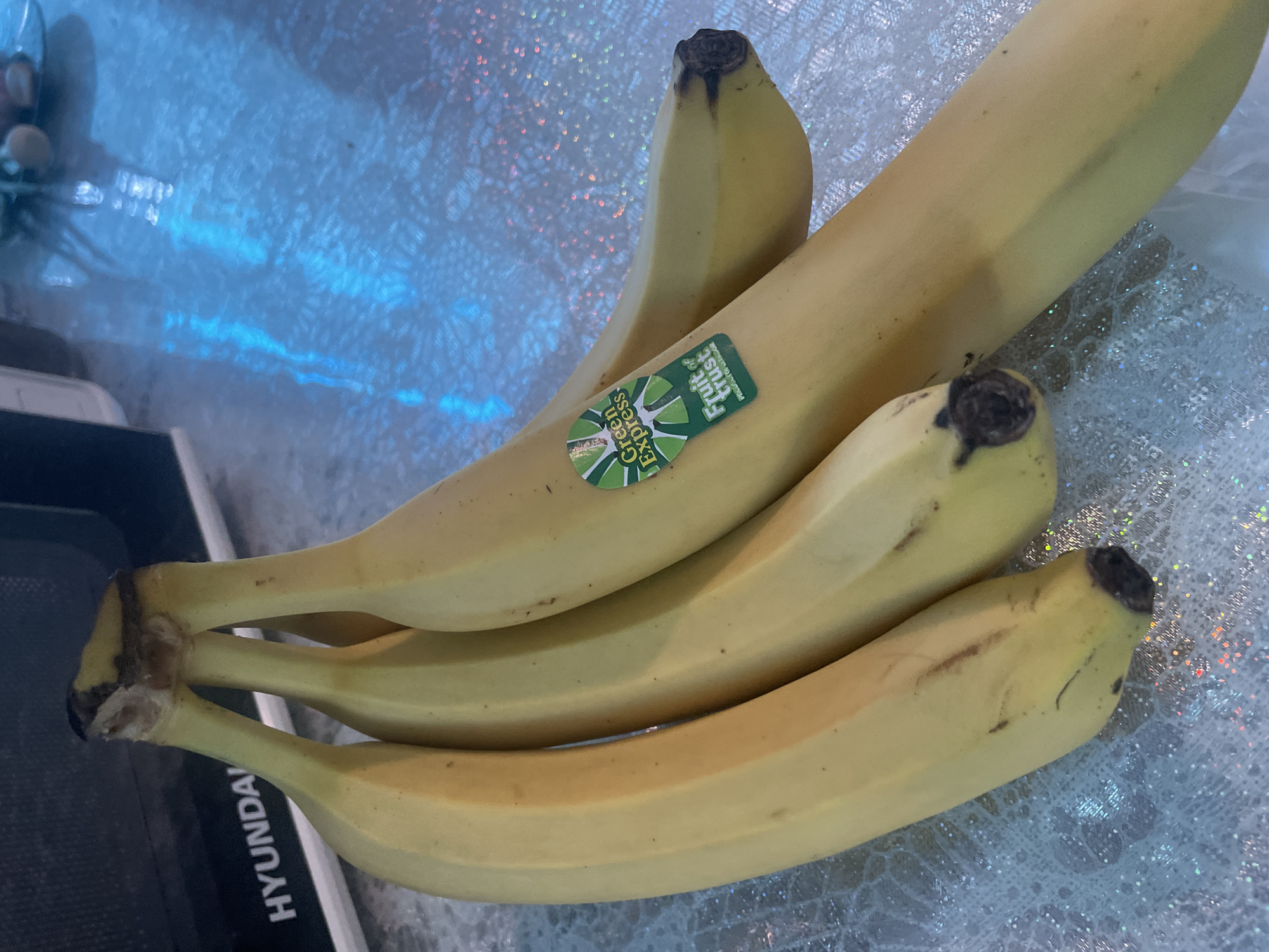 A bunch of four yellow bananas is on a glittering white and blue surface. The bananas are slightly overripe, showing brown spots and darkening at the tips. A green and white "Green Express" sticker is visible on one of the bananas. Part of a black and silver Hyundai appliance is visible in the lower left corner. The location is not depicted in the image; therefore, it is not possible to assess the location in Olginka, Russia.