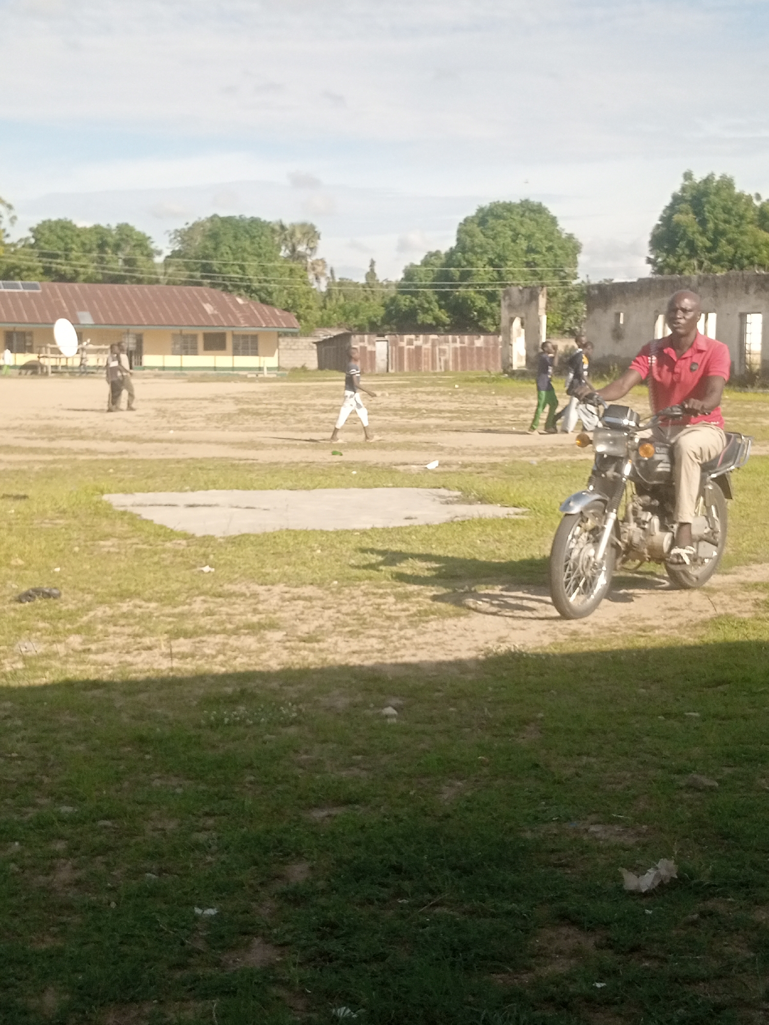 In Langtang, Nigeria, on a bright, sunny day, a man in a red polo shirt and beige pants rides a motorcycle. He is in the foreground of the image, looking to his right. The motorcycle is silver and black with a chrome exhaust. Behind him, a group of children are playing in a dusty, open field. In the background, a long, yellow building with a corrugated metal roof stands next to some partially ruined structures. Trees and a cloudy sky complete the scene. The overall atmosphere appears casual and everyday. There is no visible text in the image.