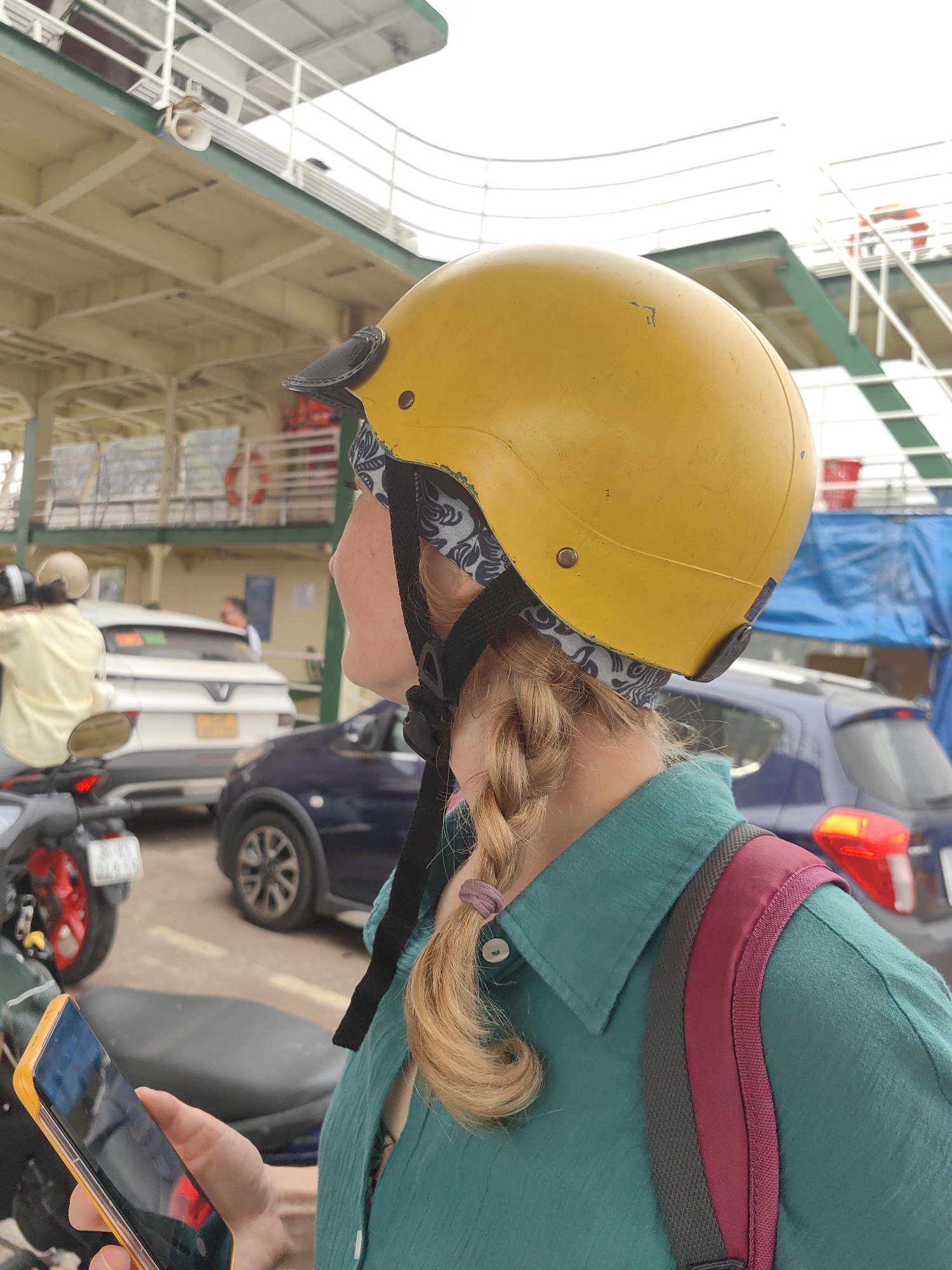 A woman with fair skin and blonde hair styled in a braid wears a yellow motorcycle helmet and a green shirt with a red and gray backpack. She holds a yellow-cased smartphone in her left hand. The backdrop features a variety of vehicles, including cars and motorbikes, suggesting a busy street or parking area. The structural components of a ship or ferry are visible overhead, suggesting a location in a port city, specifically Hải Phòng, Vietnam. The overall impression is of a traveler on a ferry, likely documenting her experience or waiting to disembark.