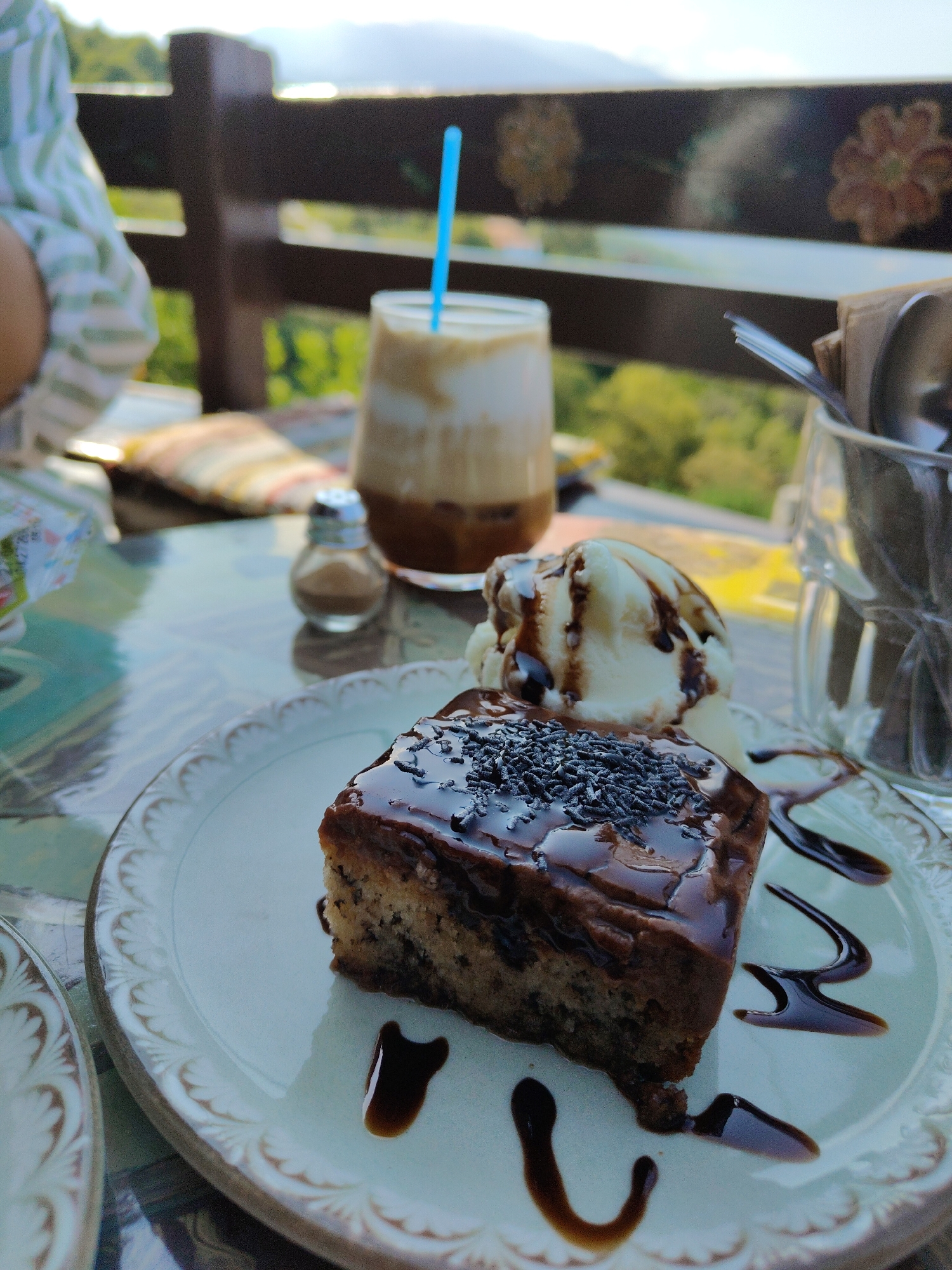 A slice of chocolate cake with dark chocolate frosting and sprinkles sits on a white plate with a decorative rim, accompanied by a scoop of vanilla ice cream drizzled with chocolate sauce. The plate is on a patterned table. In the background, a glass of iced coffee with a blue straw, a small salt shaker, and a glass container with spoons and napkins are visible. A wooden railing with decorative floral patterns is behind the table. The scene appears to be an outdoor cafe in Palaios Panteleimonas, Greece, with lush green hills and the sky in the distance.
