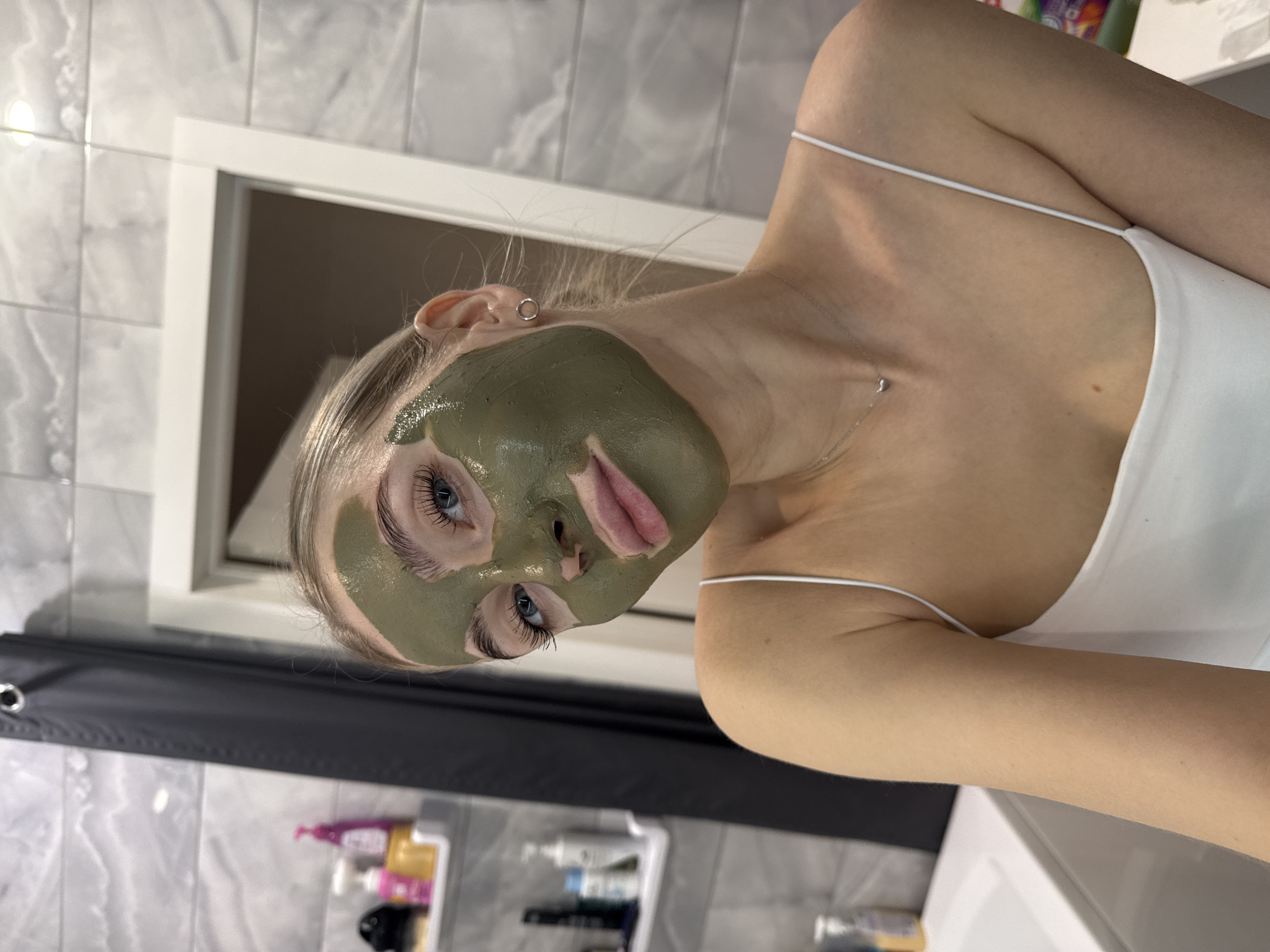 A woman is pictured indoors, likely in a bathroom based on the tiled walls and the presence of bathroom fixtures. She is wearing a white tank top and has a green facial mask applied to her face. The mask covers most of her face, leaving only her eyes and lips visible. She has blonde hair pulled back. In the background, there is a framed opening in the wall, possibly a window or mirror, and a shelf with various bottles and containers. A dark-colored shower curtain is also visible. The image's angle suggests it might be a selfie or a photo taken at an unusual perspective.
There is no information suggesting this image was taken in Izhevsk, Russia.