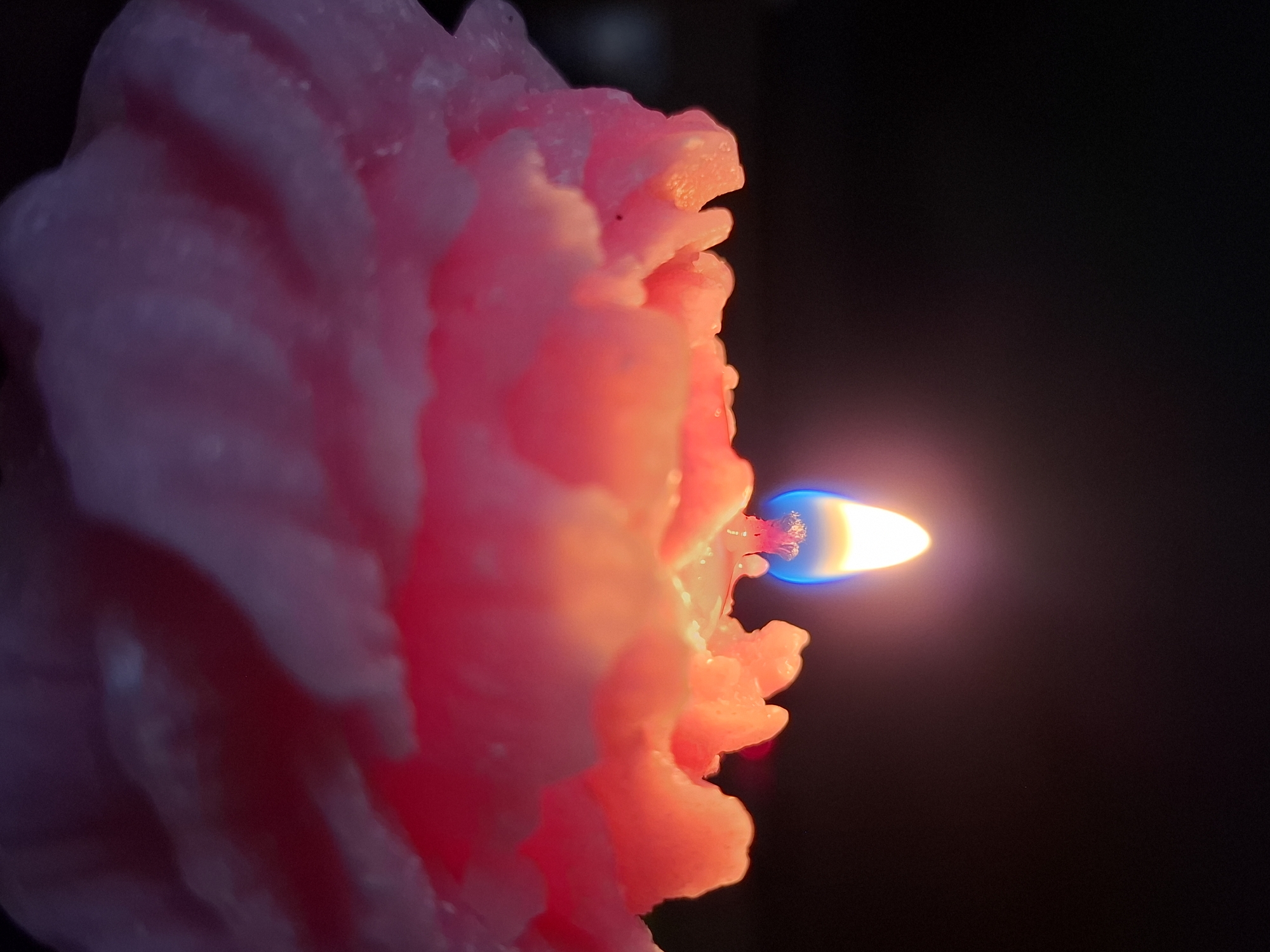 A close-up, vertical shot features a vibrant, burning pink or coral-colored candle against a stark, dark background. The candle, which occupies the left side of the frame, has a textured, possibly ruffled or ridged surface, catching the light from its own flame. The wax closest to the flame glows with warm orange and red hues, highlighting its intricate details and melted edges.
The candle's flame, positioned on the right, is a prominent focal point. It exhibits a distinct structure: a bright, almost white-yellow core, surrounded by an orange-yellow body, and a vivid blue base where it emerges from the wick. The flame casts a warm, soft glow onto the surrounding wax, creating a stark contrast with the deep black background which offers no discernible details or objects.
The overall impression is one of warmth, intimacy, and tranquility, emphasized by the sole light source in the darkness. The time of day appears to be night or an indoor setting with very low ambient light. No specific location cues are visible within the image itself, but the context provided is Chaikovsky, Russia. There is no visible text in the image.