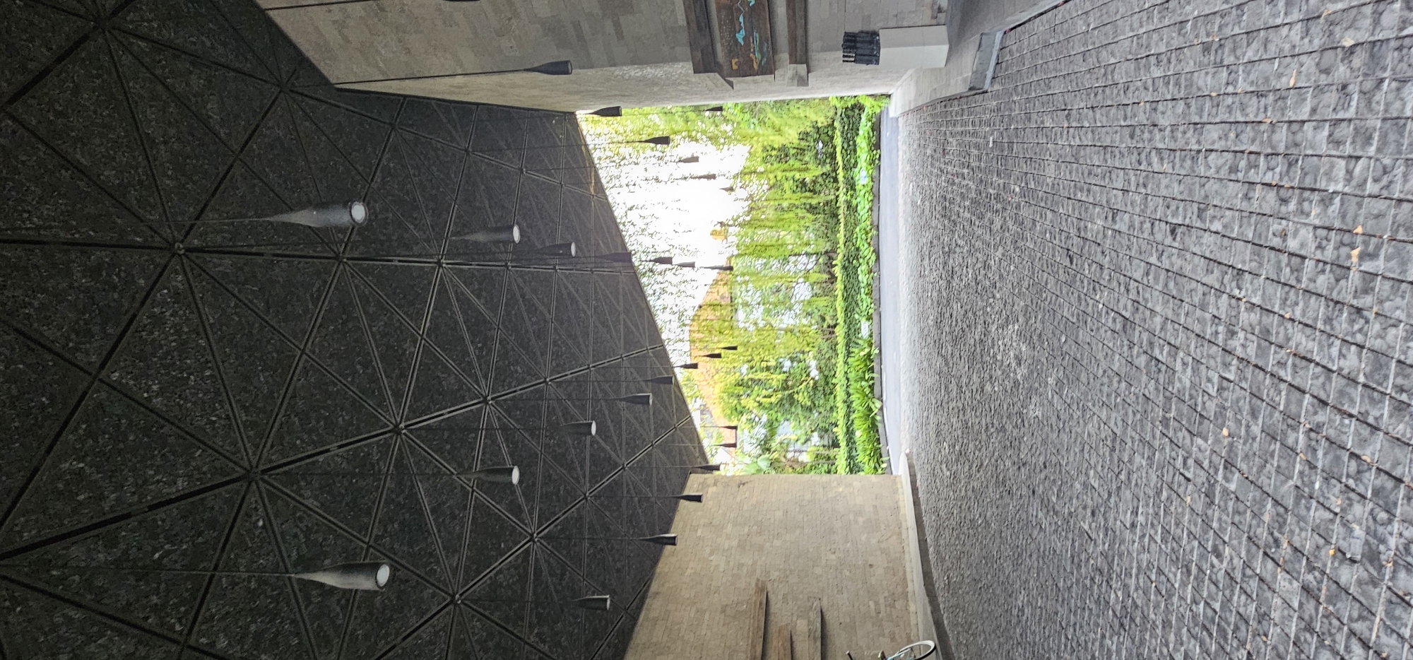 The image depicts an architectural view, likely a covered pathway or entrance area in Denpasar, Indonesia. The left side features a wall with a striking geometric pattern of triangles, adorned with numerous conical light fixtures extending outward. A glimpse of lush greenery, including trees and possibly vines, is visible through the opening. The right side showcases a textured, brick-like pathway leading away from the viewpoint. The ground is paved with dark, rough-hewn stones. The perspective is slightly tilted, lending a dynamic feel to the composition. A small portion of a bicycle is also visible near the bottom of the image.
