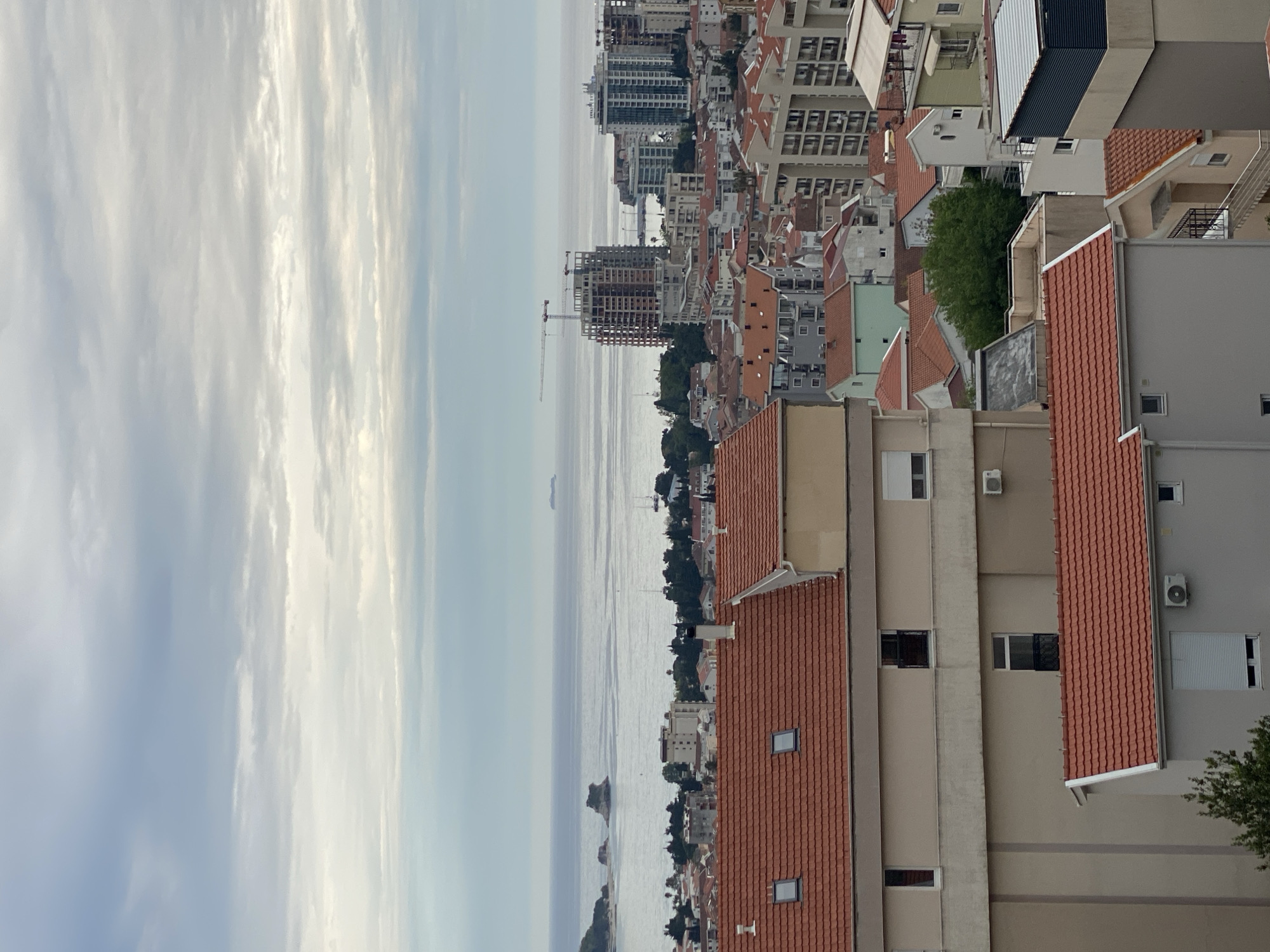 The image captures an aerial view of Budva, Montenegro, featuring the city's architecture, the Adriatic Sea, and the sky. The city is densely populated with buildings, many of which have orange or red-tiled roofs. High-rise buildings and construction sites are visible in the distance. The sea occupies the left side of the frame, with a few islands and a visible horizon. The sky above is cloudy with varying shades of blue and white. There are no people or discernible actions visible in the image.