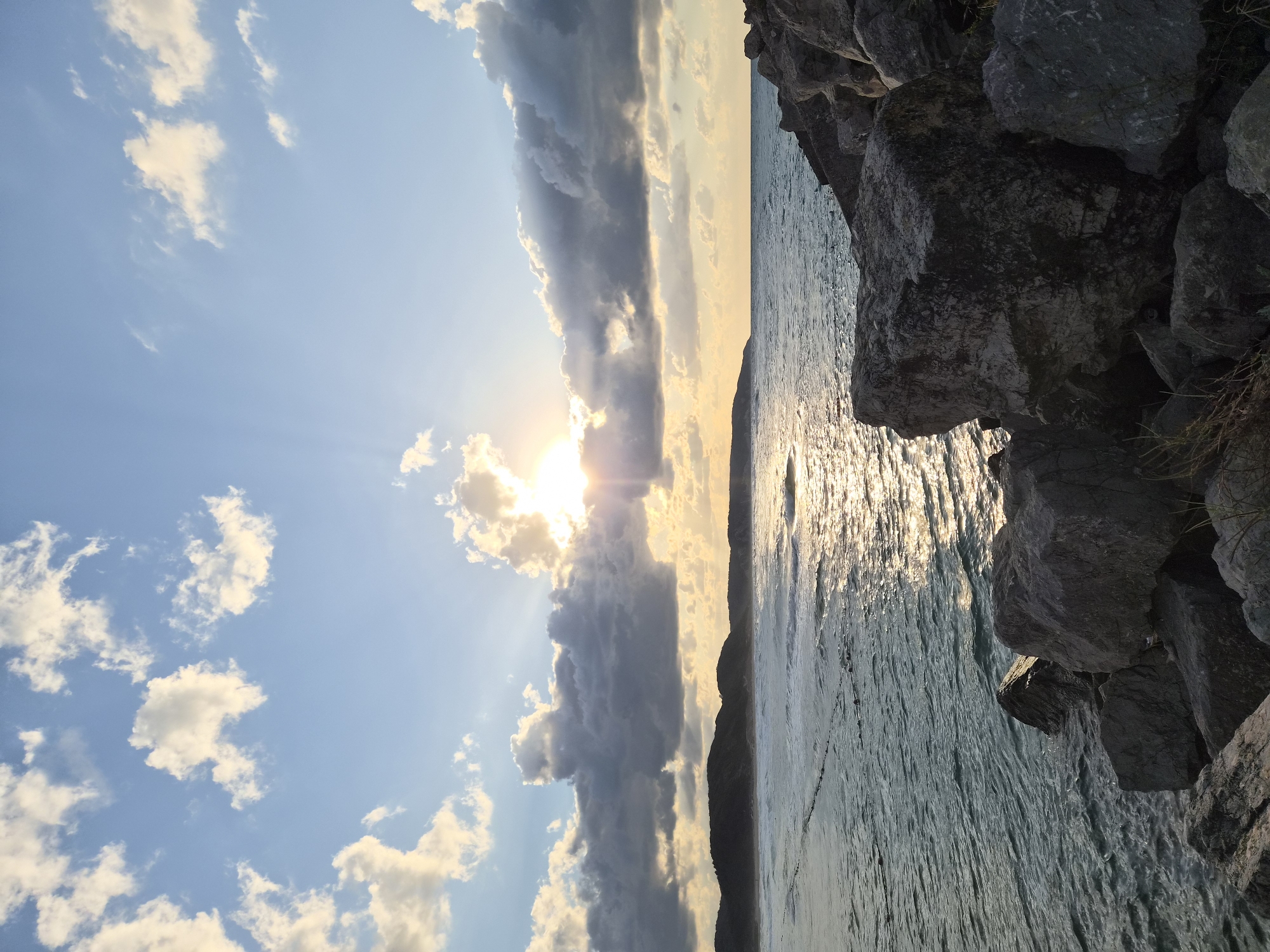 A vertical, eye-level, medium-wide shot captures a sunset over the Black Sea at the Şile coast in Turkey. The sun, a bright orb partially obscured by clouds, casts a golden glow across the sky and water. Rays of light emanate from the sun, piercing through the clouds and reflecting on the rippling surface of the sea. The water is a deep, tranquil blue, with waves gently lapping at the shore. In the foreground, large, rough-hewn rocks, some covered in moss and grass, line the coastline, creating a natural barrier between the land and the sea. In the distance, a range of low, verdant hills forms the backdrop to the scene, their silhouettes softened by the twilight haze. The sky is a canvas of blue, dotted with fluffy white and dark grey clouds, adding depth and texture to the celestial panorama. The overall mood is one of serenity and natural beauty, with the vibrant colors of the sunset creating a breathtaking spectacle.