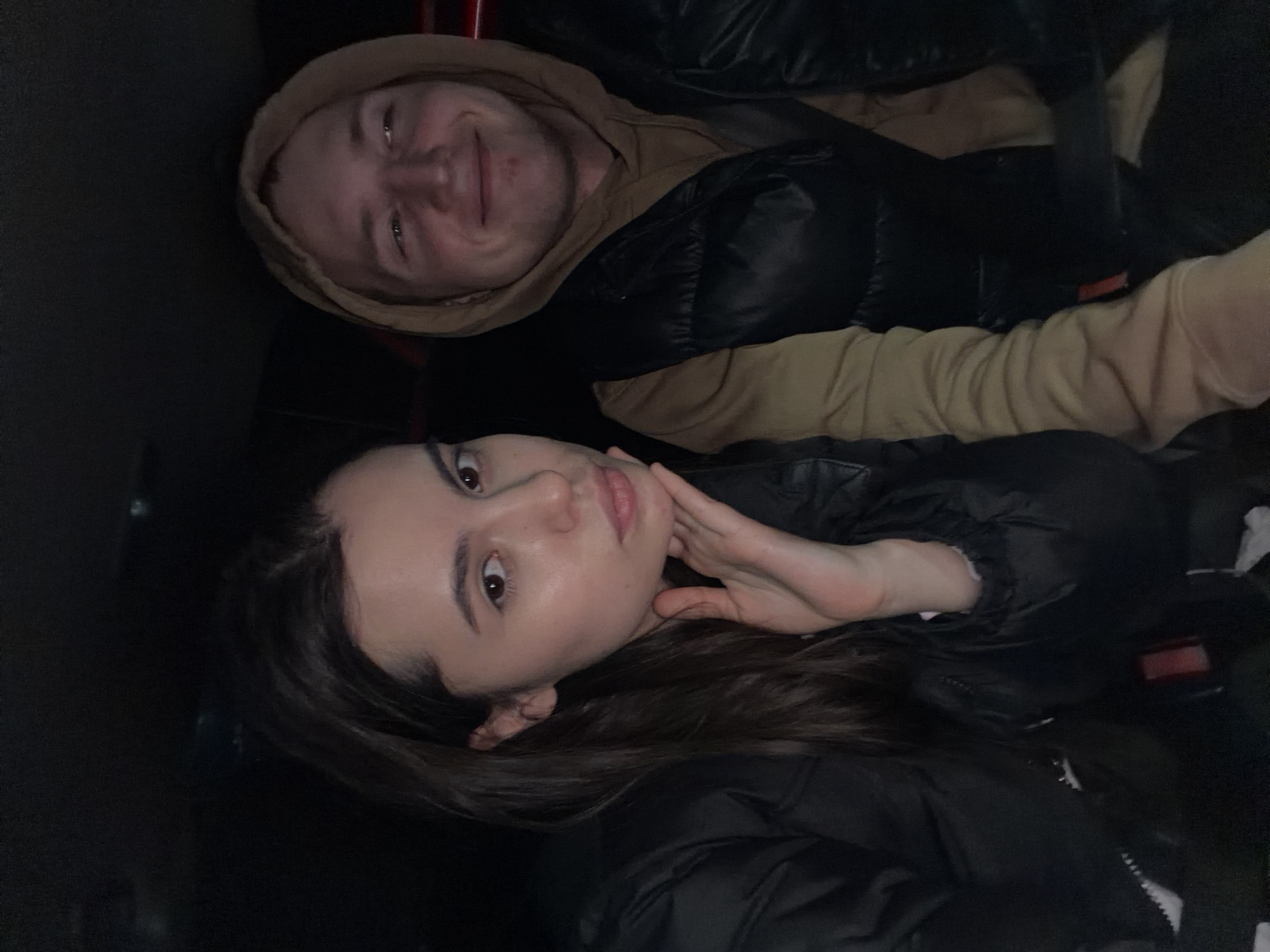 This vertical-format photograph captures two individuals, a man and a woman, in what appears to be a dimly lit interior space, likely a car, based on the dark, confined surroundings and hints of red interior elements. The scene takes place in Chaikovsky, Russia.
The man, positioned in the upper part of the frame, has light hair and is smiling directly at the camera. He wears a light brown hooded sweatshirt underneath a dark, possibly black, puffer jacket.
In the foreground, the woman looks directly at the camera with a neutral, poised expression. She has long, dark hair, dark eyes, and wears a dark, possibly black, puffer jacket. Her left hand is gently placed against her jawline.
The overall atmosphere is casual and intimate, with both subjects seemingly posing for the photograph. The low light conditions suggest it could be evening, night, or an indoor setting. There is no visible text in the image.