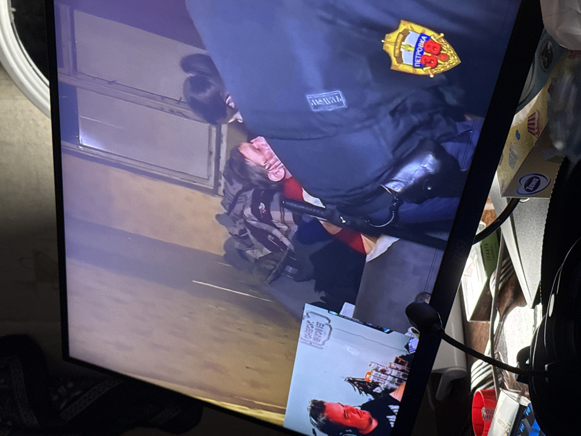 The image depicts a scene on a monitor screen, showing an individual in what appears to be a police uniform with a badge that says "ПЕТРОВКА" (Petrovka) in Cyrillic, which is a well-known police precinct in Moscow, Russia. This officer seems to be restraining another individual who is lying down. There are boxes next to the monitor, and part of a headset can be seen on the right side of the image.