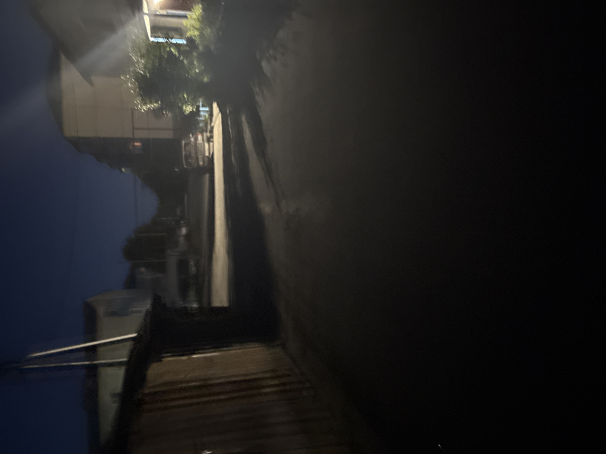 This nocturnal image depicts a narrow, unpaved road or alleyway in an urban or suburban setting. On the left, a dark, single-story structure with vertical paneling, possibly a garage or shed, stands adjacent to the road. A utility pole with multiple wires extends upwards near this structure. Further down the left side, several light-colored, multi-story buildings are visible against the dark sky. In the midground, an older model, light-colored vehicle, possibly a pickup truck or SUV, is parked on the right side of the road, partially illuminated. On the immediate right, a prominent multi-story building with a light-colored facade is present. A bright artificial light source, emanating from this building, casts strong illumination onto the surrounding area, including dense green foliage, and creates a distinct lens flare effect extending into the frame. Shadows are prominently cast across the road and ground due to these light sources. The sky is dark blue-black, indicating nighttime. No individuals are visible.