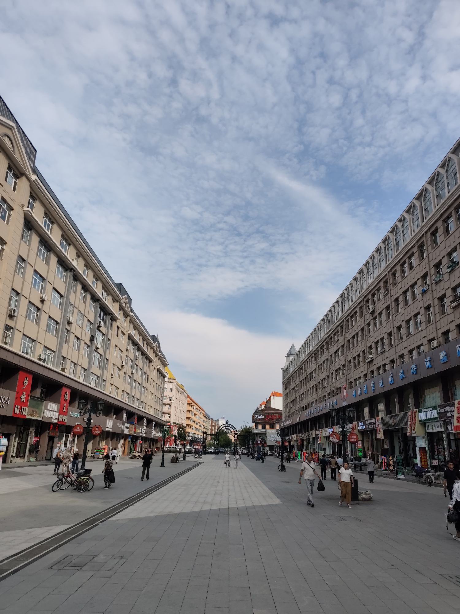 The image captures a street scene in Aihui, China, with two large, multi-story buildings lining either side of a wide pedestrian walkway. The buildings exhibit a European architectural style with numerous windows and decorative elements. The walkway is paved with gray tiles and features some dark inlaid lines. Several people are walking and standing on the walkway. On the left side, a bicycle is parked. Signs with Chinese characters are visible on the storefronts at the ground level of both buildings. The sky above is blue with scattered white clouds. The overall impression is a bustling urban environment on a clear day.