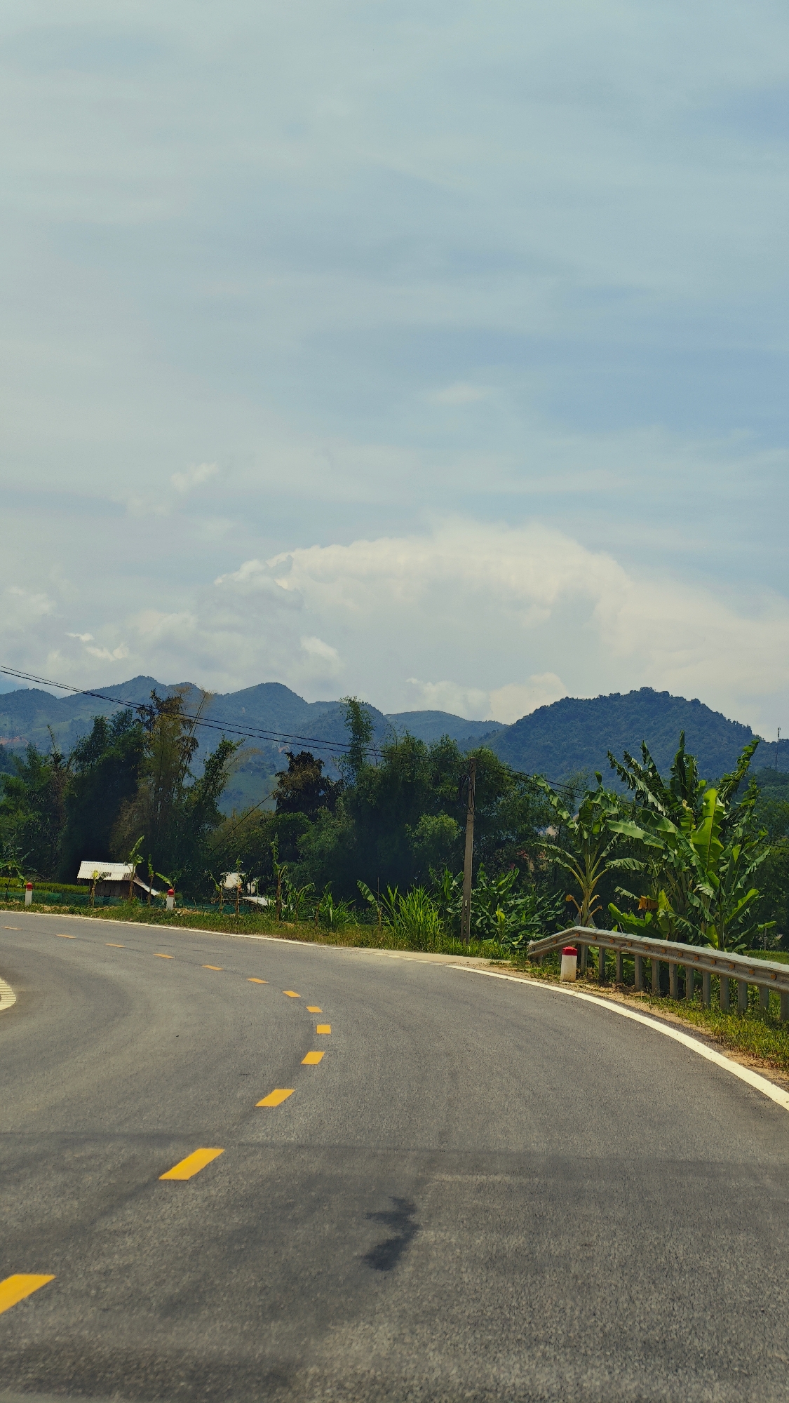 A scenic, wide-angle shot captures a winding asphalt road cutting through a verdant, mountainous landscape in Duong Quy, Vietnam. The road, devoid of vehicles or people, features distinct dashed yellow lines marking its center and a solid white line defining its right edge. On the right side, a metal guardrail with a prominent red and white marker post runs alongside the road, separating it from dense tropical foliage, including large banana plants and various trees.
To the left of the road, more lush green vegetation and tall trees create a dense boundary. Peeking through the foliage, a small, rustic structure with a light-colored roof is visible, hinting at local dwellings. A utility pole with power lines stretches across the midground, adding a touch of infrastructure to the natural scenery.
In the background, multiple layers of majestic mountains rise, their peaks shrouded in a soft haze, suggesting distance and scale. The sky above is partly cloudy, with scattered white cumulus clouds against a light blue expanse, indicating a bright daytime setting, likely late morning or early afternoon. Shadows on the road surface are subtle, further supporting the time of day. The overall atmosphere is tranquil and inviting, showcasing the natural beauty of the region. No visible text is present in the image.