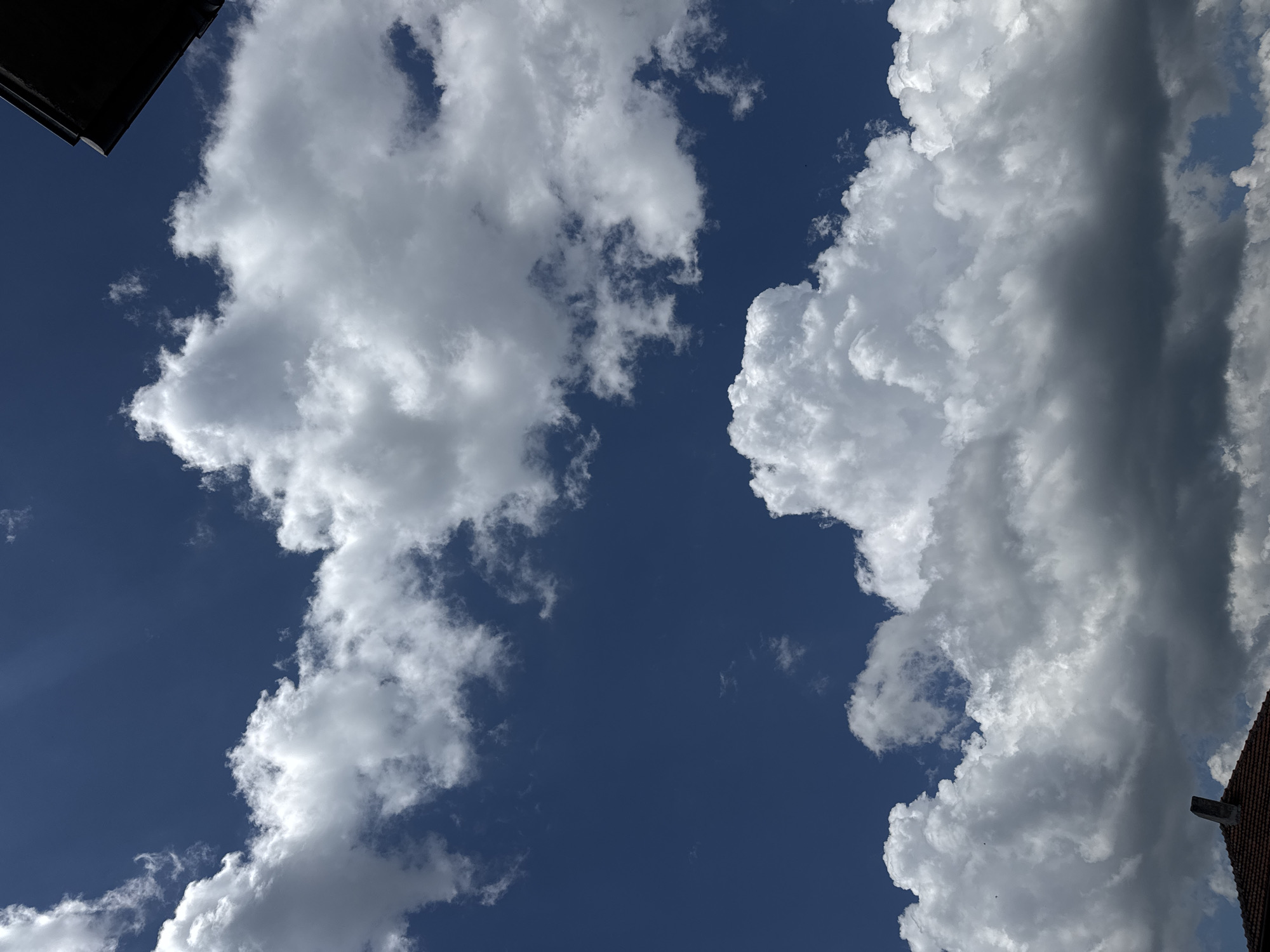 The image shows a view of the sky in Sarnevo, Bulgaria. Two large clusters of white, fluffy cumulus clouds are visible against a deep blue sky. The corner of a dark building is visible in the top left corner of the frame, and a section of a red-tiled roof is visible in the bottom right corner. The image is shot from a low angle, looking upwards. No people or actions are visible. The environment is a clear, sunny day.