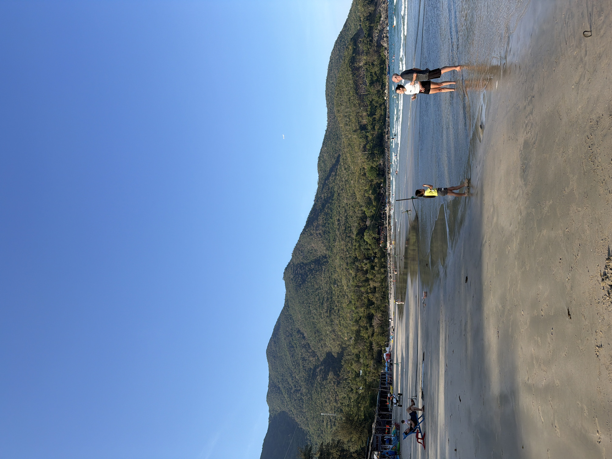 The image depicts a beach scene in Nam Nha Trang Ward, Vietnam, with a tall, forested mountain forming the backdrop. A clear, blue sky is overhead. In the foreground, the sandy beach is visible with a couple standing in shallow water near the shoreline. A single child stands further down the beach, also in shallow water, near what appears to be a fishing rod. Beach chairs and sunshades are set up along the edge of the beach, occupied by other people. The water is clear and reflects the sky.
