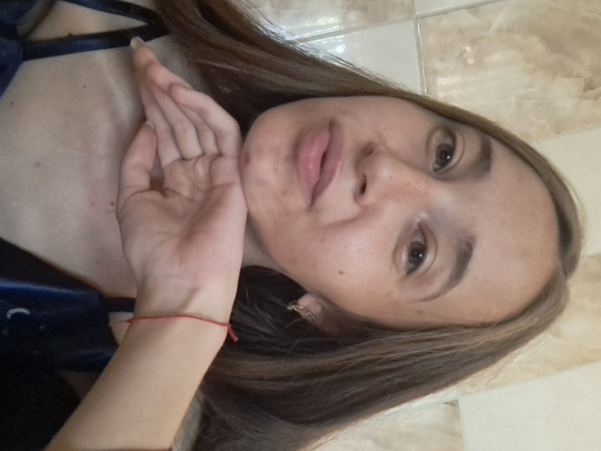 A young woman with long brown hair is pictured in a selfie, resting her chin on her left hand. She is wearing a dark blue garment with thin straps. A red string bracelet is visible on her left wrist. The background appears to be a tiled wall. The image was taken in the city of Vypasne, Ukraine.