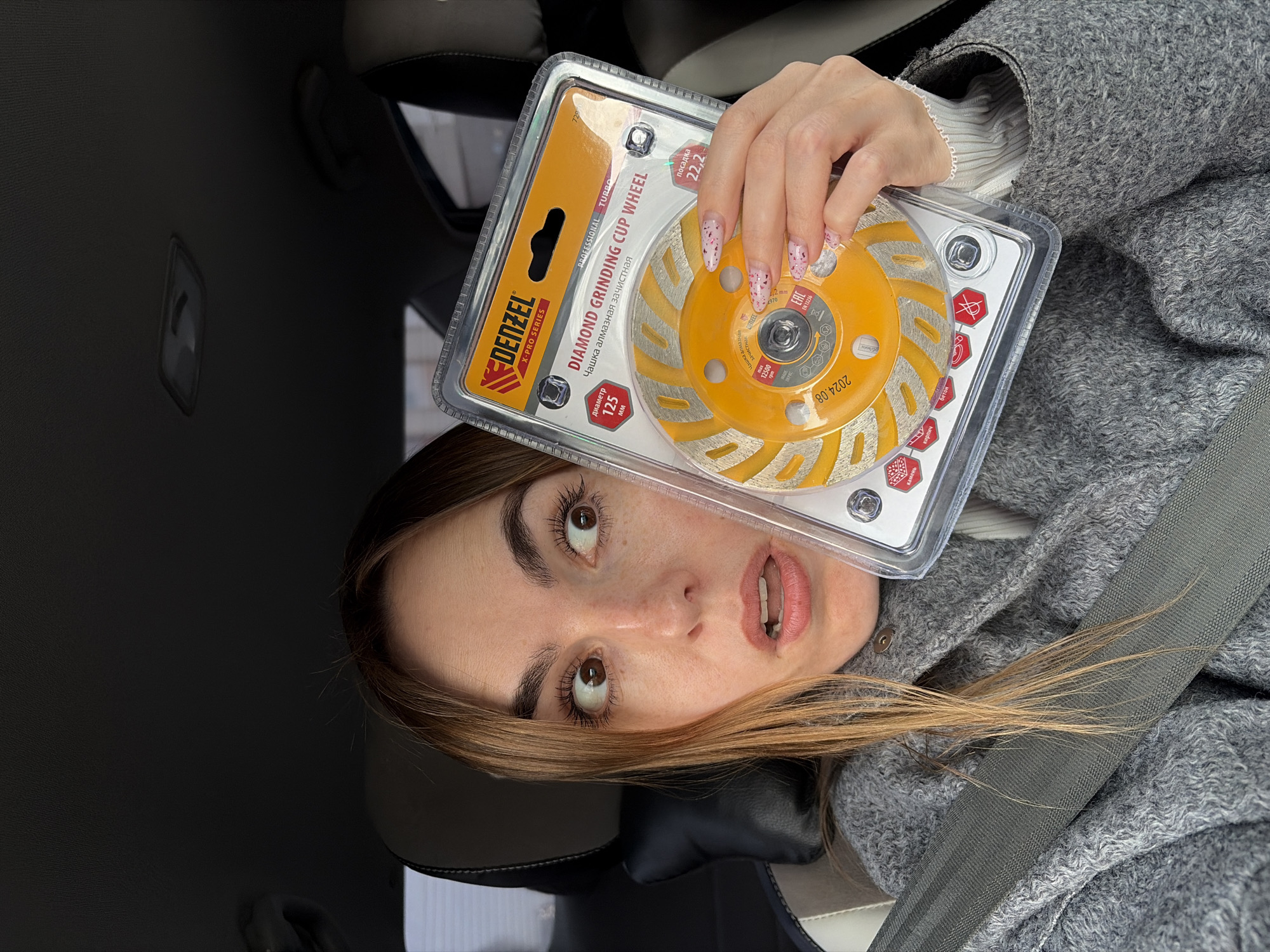 The image shows a woman indoors, possibly in a car, holding a "DENZEL X-PRO SERIES" DIAMOND GRINDING CUP WHEEL in its packaging. The woman has brown hair and is wearing a gray coat or sweater and a white long-sleeved shirt. She is wearing a seatbelt. Text on the packaging indicates "диаметр 125 MM" and "посадка 22,2." The text on the diamond grinding wheel is in Russian. The location is not provided and therefore is unknown.