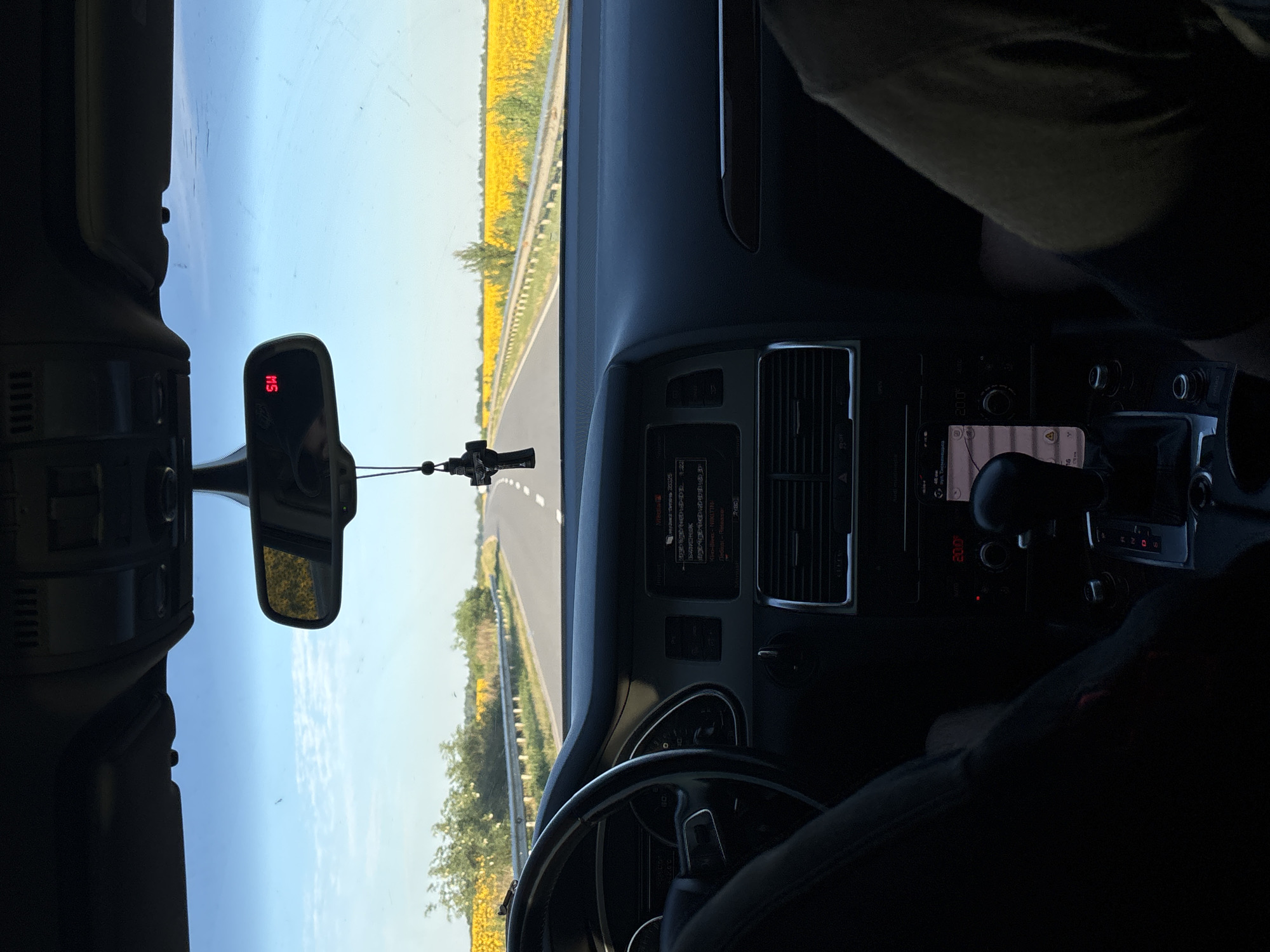 The image is taken from inside a car, looking forward through the windshield. The car is on a road that passes through a field of sunflowers. The sky is blue with some scattered clouds. On the dashboard, there is a navigation screen showing a map and route. A car accessory, a cross, is hanging from the rearview mirror. The road appears to be in a rural area, possibly outside the city of Buchach, Ukraine, given the presence of sunflower fields which are common in the region.