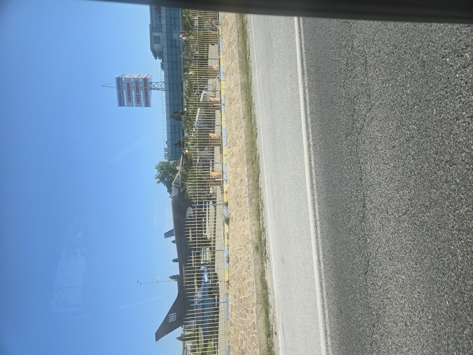 The image depicts a roadside scene in the city of Unknown, Unknown, captured on a bright, sunny day. A two-lane road stretches along the right side of the frame, composed of dark asphalt. Adjacent to the road, a low stone wall borders a grassy area. A military aircraft, possibly an old fighter jet or transport plane, is displayed behind a metal fence. The aircraft is painted in dark colors and has the letters "BH" visible on its tail fin. In the background, there is a tall building with large windows. A sign stands in front of the building. The image seems to have been taken from inside a vehicle moving along the road.
