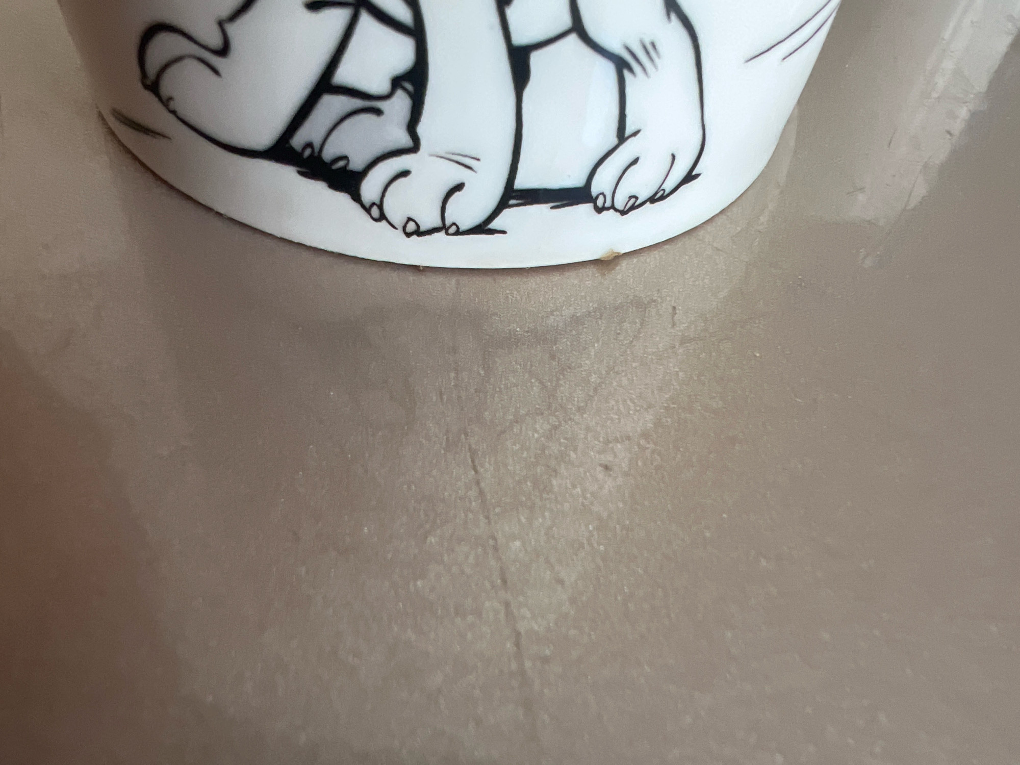 A close-up, low-angle shot shows the bottom portion of a white ceramic mug decorated with black line drawings of a dog's paws. The mug rests on a smooth, reflective surface that appears to be a countertop or table, with a neutral, muted tone. The surface shows some minor smudges and reflections, indicating it is likely clean but not pristine. The dog's paws are depicted in a cartoonish style, with rounded toes and simple line details for claws and paw pads. The lighting is soft and even, casting no strong shadows. The background is a blurred continuation of the reflective surface, providing no additional context about the environment or location.