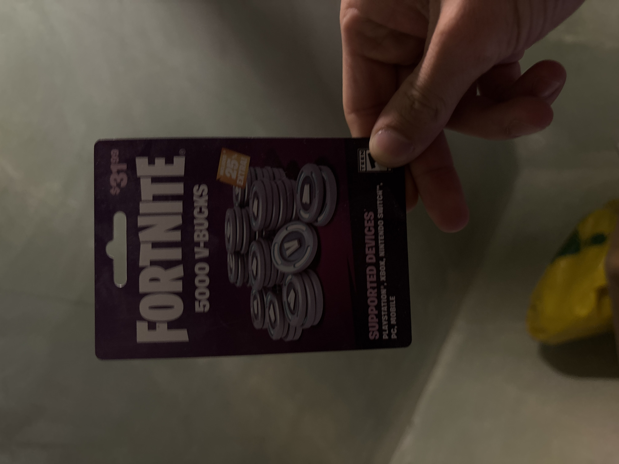 A hand holds a "Fortnite" V-Bucks gift card. The card is purple and advertises 5000 V-Bucks for $31.99. The card also mentions that the V-Bucks are compatible with PlayStation, Xbox, Nintendo Switch, PC, and mobile devices. In the background, there is a white surface and a yellow object.