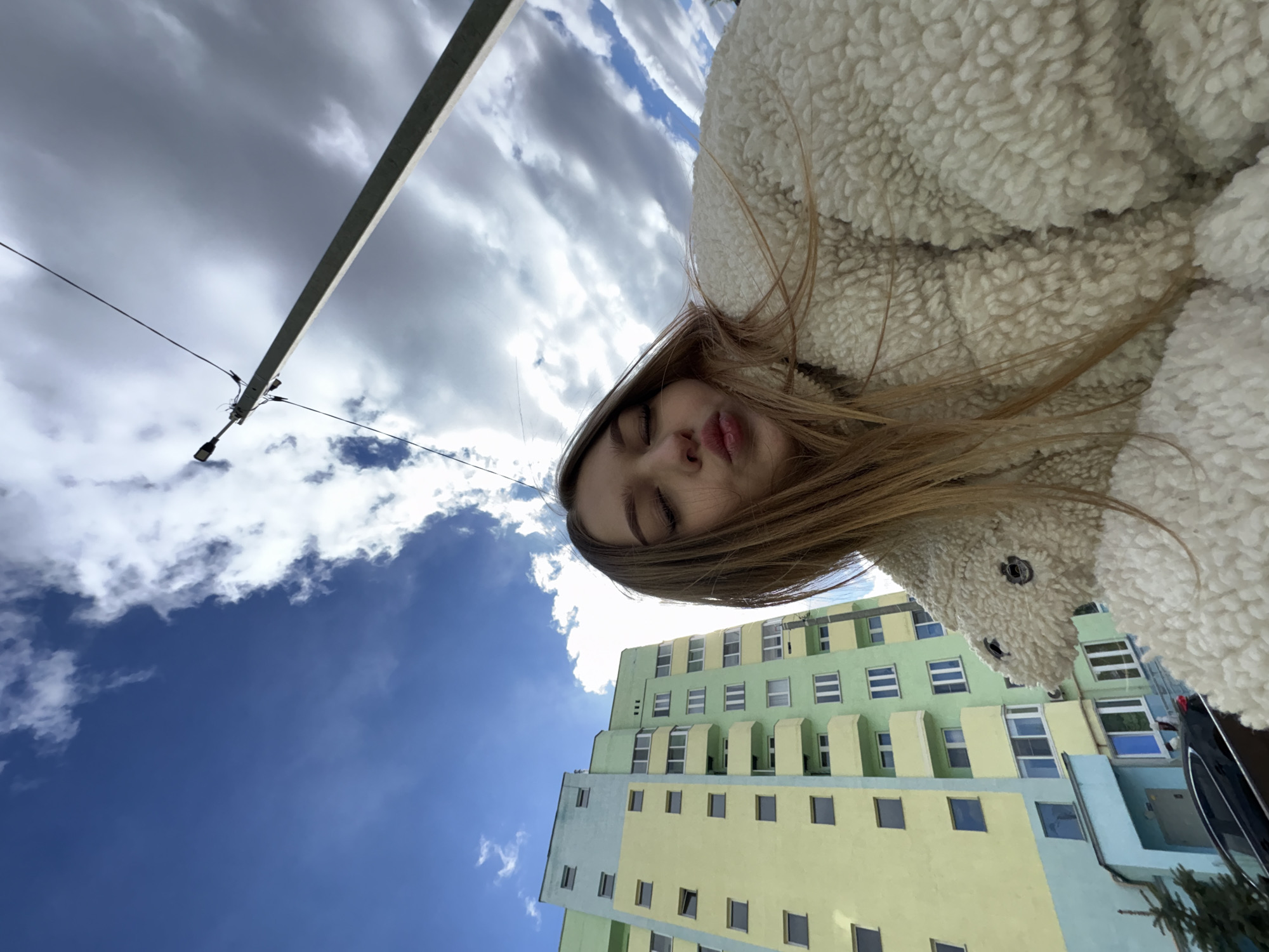 A rotated low-angle shot shows a young woman in a fluffy white jacket. She has fair skin and light brown hair, and her eyes are closed. Her mouth is slightly pursed. In the background is a multi-story building painted in pastel yellow and green, typical of post-Soviet architecture. The sky is partly cloudy with both dark and bright patches. A utility pole is visible in the upper left corner. The location appears to be in a city, potentially Chaikovsky, Russia, based on architectural styles common in the region.