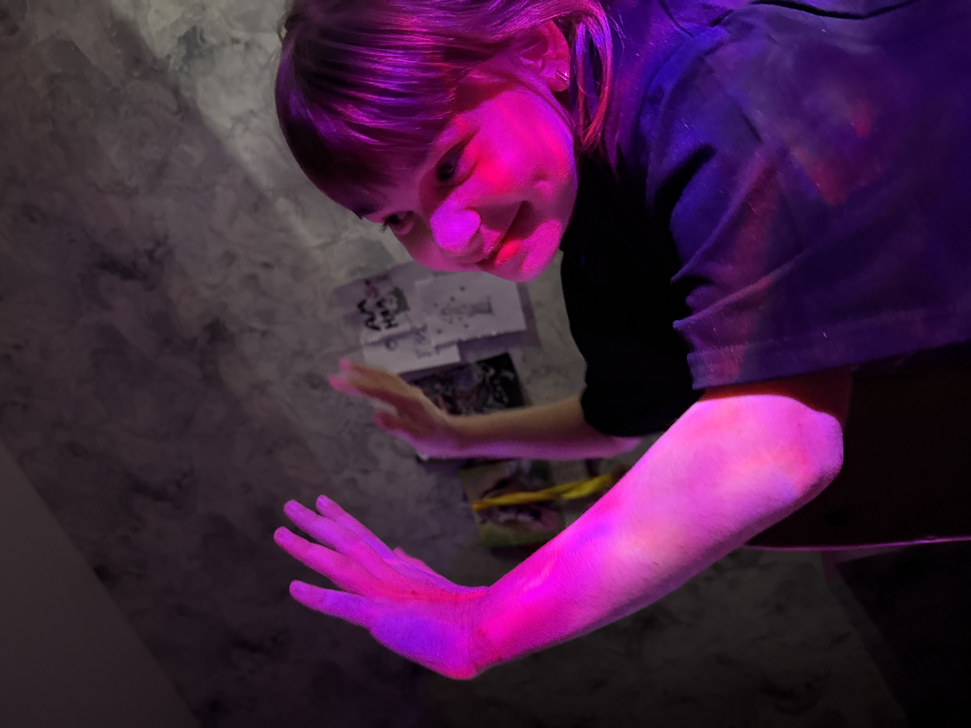 A person, possibly a child, is under intense pink and purple lighting, casting the scene in a surreal glow. The person's face is illuminated with a bright pink hue, while their hair appears purple. The subject seems to be interacting with objects on a surface, possibly a table, where items such as cards or papers are visible. The background features a textured wall with a marbled effect. The specific location is not discernible from the image. The primary action involves the person's attention focused downward on the objects.