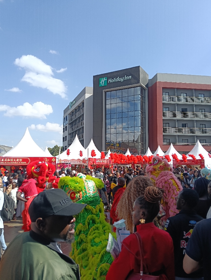 An outdoor cultural festival is depicted in Nairobi, Kenya, identifiable by the modern, multi-story Holiday Inn Express building visible in the background, situated on Thika Road. The sky is clear blue with scattered white clouds. Numerous white pop-up tents, adorned with red lanterns and banners displaying Chinese characters, including "Embassy of the People's Republic of China in Kenya," line an open area. A large crowd of people, predominantly of African descent, fills the foreground and midground. Several performers in vibrant lion dance costumes—specifically a green, a red, and a pink/yellow one—are moving through the gathering, indicating a performance or parade. In the immediate foreground, the back and right profile of a man wearing a black baseball cap and a dark green jacket is visible. To his right, a woman with intricately styled brown hair and a red top is seen from behind, wearing a gold-colored earring. Attendees are observing the festive activities.