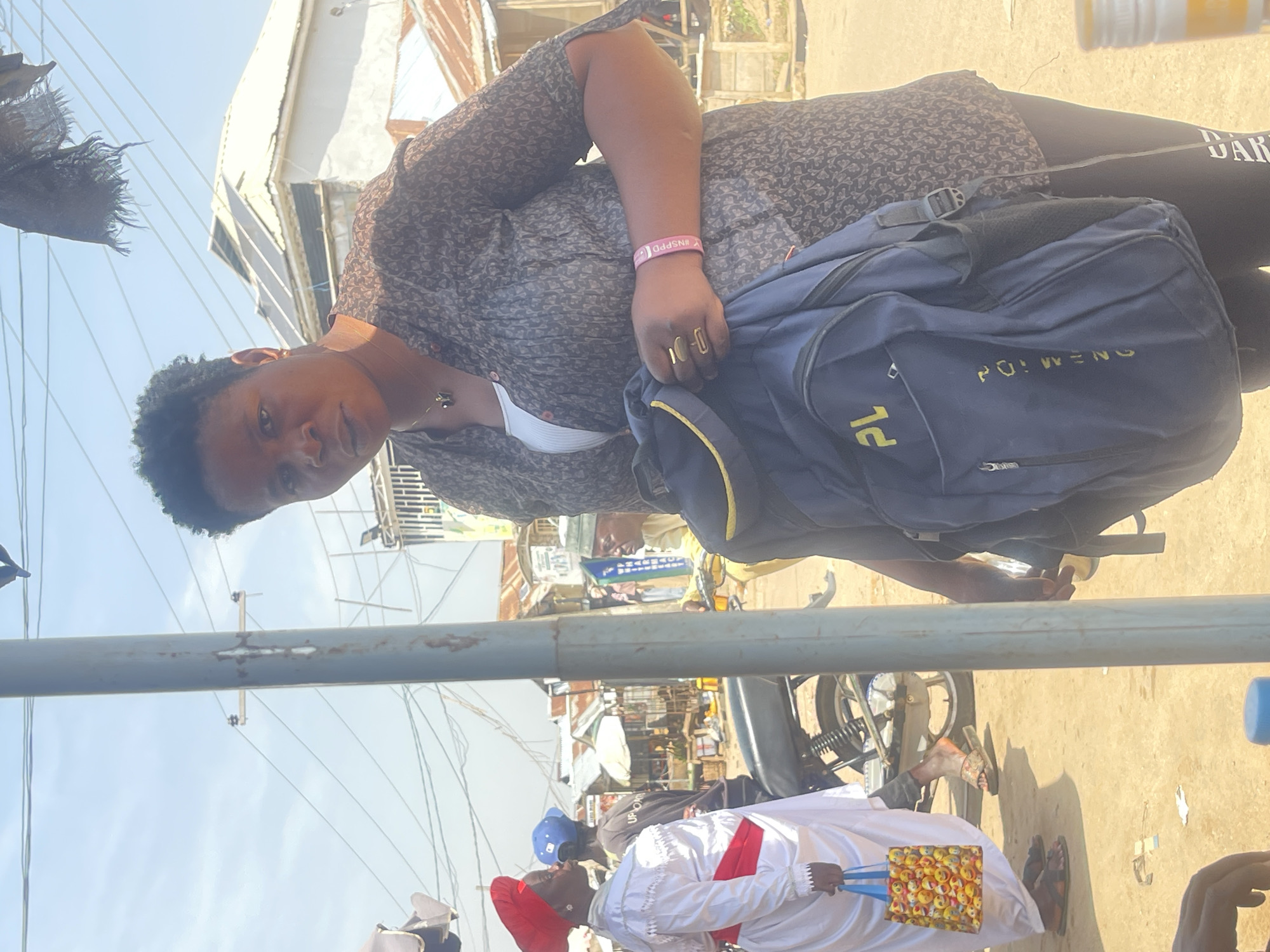 In the city of Abuja, Nigeria, a medium shot captures a woman standing in front of a light pole. She is wearing a patterned brown shirt over a white undershirt and dark leggings, holding a blue backpack. Her hair is short and black. On her left wrist, she wears a pink bracelet. A gold ring is on her right index finger. To her left, a person in a white robe with a red sash and a red head wrap is holding a colorful bag filled with emoji faces. Behind them, a motorcycle is parked. The background shows a street scene with buildings, power lines, and a clear sky, suggesting an outdoor market or thoroughfare. The overall lighting indicates a bright, sunny day.