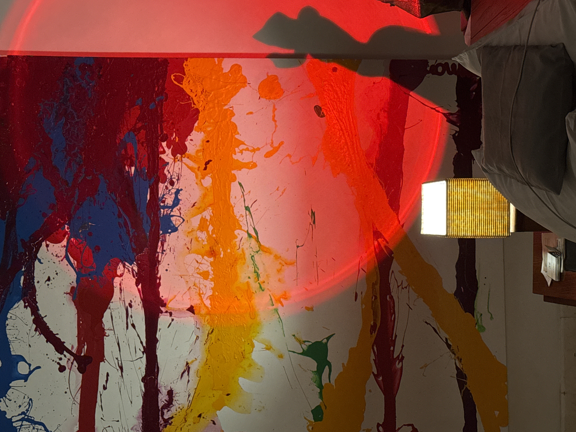 The image captures an indoor scene, likely a bedroom, featuring a vibrant abstract painting illuminated by artificial light. The painting, oriented vertically, displays a dynamic splatter and drip style with prominent strokes of red, blue, yellow, orange, and hints of green and dark red/purple paint on a white background.
A striking, large circular beam of intense red light is projected across the upper and middle sections of the painting and the wall above it, creating a dramatic atmosphere. An irregular, dark shadow is cast on the wall in the upper right, interacting with the red light. To the far right, a small, rectangular lamp with a ribbed, possibly wooden or wicker shade emits a warm, yellowish light, casting localized illumination.
On the right side of the frame, the edge of a bed with a white sheet and a grey pillow is visible, suggesting the painting is hung in a sleeping area. A small shelf or bedside table next to the bed holds a few indistinct white and dark objects. The interplay of the painting's bright colors with the red ambient light and the warm glow of the lamp indicates an evening or nighttime setting. Faint, dark letters, possibly forming a word like "flow," are subtly integrated into the paintwork on the lower right section of the painting, partially obscured by the red light. The overall mood is artistic and contemplative.