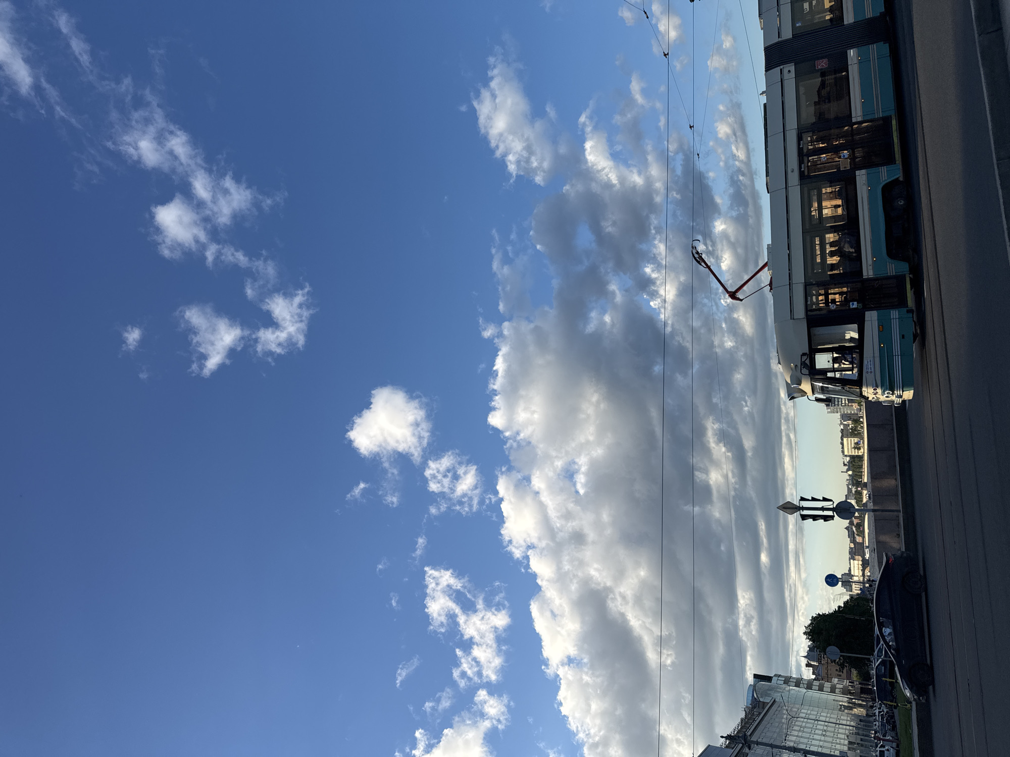 The image depicts a cityscape in Saint Petersburg, Russia, under a bright blue sky with scattered white clouds. A modern tram, painted in teal and white, travels along its tracks in the foreground, its pantograph extended to connect with overhead wires. To the left, a black sedan is parked on the side of the road, and beyond it, a contemporary building with a glass facade stands tall. Further in the distance, a skyline of buildings is visible, with a body of water suggesting a canal or river. Traffic lights and street signs are present on the sidewalk. The overall scene is characteristic of an urban environment during the daytime, with signs of public transportation and daily life.