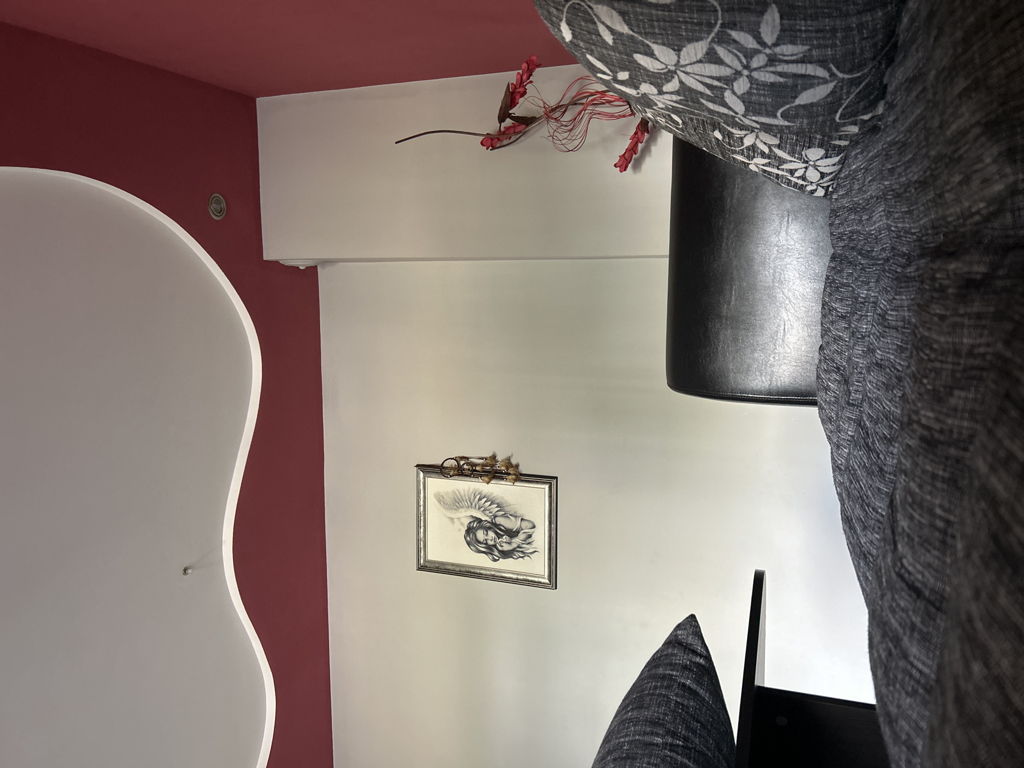 The image displays an interior room, likely a living room or lounge, with a distinctive modern design. The ceiling is painted a deep red (maroon), contrasting sharply with the white walls. On the left side, a flowing, curved white architectural feature accentuates the wall, near which a small recessed light is visible.
Centered on a white wall, a framed piece of art depicts a grayscale drawing of an angelic figure with large wings, appearing to be in a serene, sleeping, or meditative pose. Above the frame, a small, ornate decoration, possibly a brass horn or trumpet adorned with feathers, is mounted.
To the right, a white horizontal surface, perhaps a shelf or part of the ceiling design, hosts a decorative branch with artificial red flowers or berries and slender red wire-like elements.
In the foreground and to the right, a dark-colored sofa or chair is prominent. It has a black, leather-like base and is accessorized with two cushions: one featuring a white floral or leafy pattern against a dark grey background, and another solid dark grey cushion. The overall scene suggests a contemporary and artfully decorated space. There is no visible text in the image.