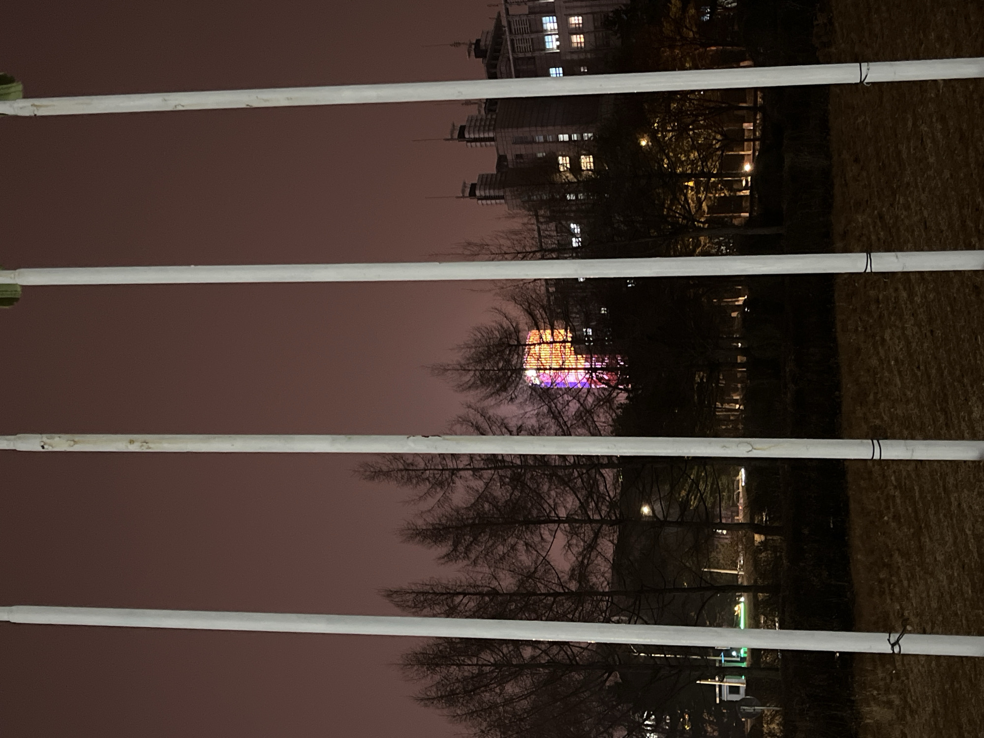 The image, shot at night in Changchun City, China, shows a view through a fence of white horizontal bars. In the background are buildings with illuminated windows and structures, obscured by leafless trees. The sky is dark and overcast. The scene is lit by artificial lights from the city.
