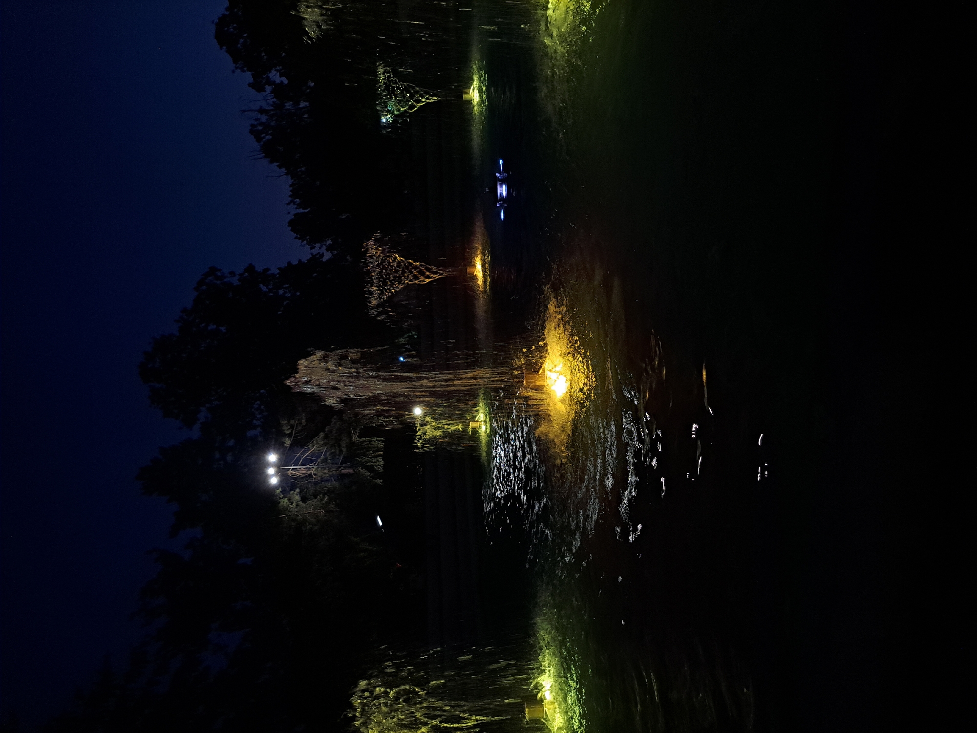 This is a nighttime photograph of a water feature with artificial lighting. The scene appears to be a park or garden area with several illuminated fountains and cascading water. The water is lit with yellow and green lights, creating a dramatic effect against the dark sky and silhouettes of trees. In the center of the frame, a fountain is spraying water upwards, illuminated by a bright yellow light at its base. To the right, another fountain has a more textured, cone-like spray, also lit from below. In the distance, a small, blue-lit object floats on the water. Several spotlights are visible in the background, illuminating the trees and the water. The overall impression is one of tranquility and ethereal beauty, enhanced by the play of light and water in the darkness. The location is identified as being in Popovo, Bulgaria. There are no people visible in the image.