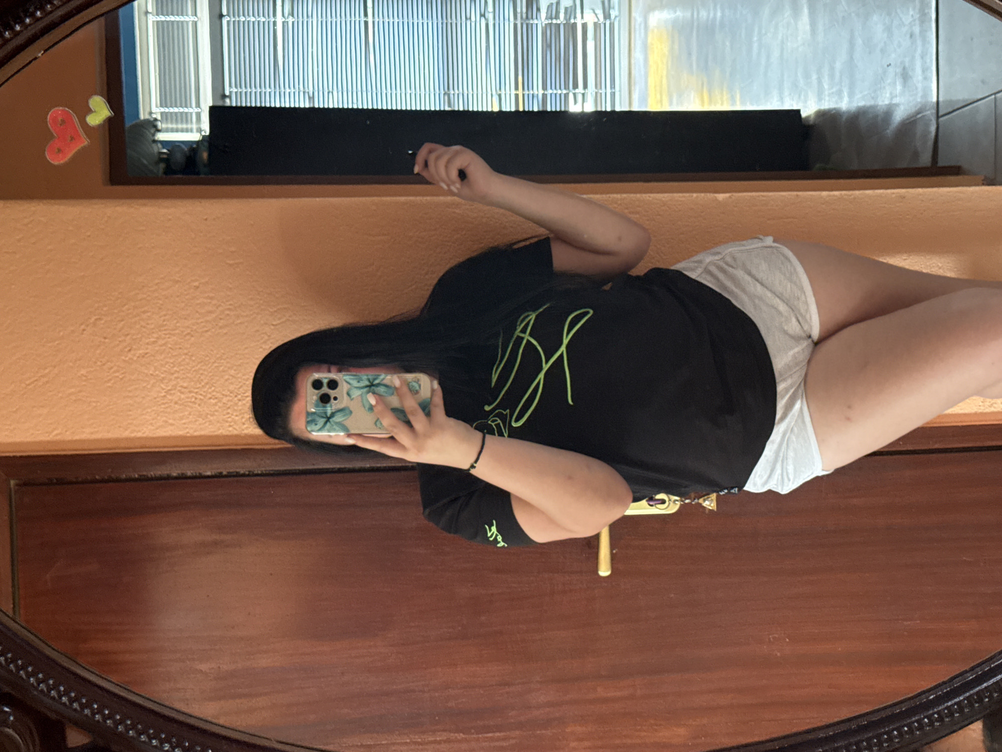 An individual is captured in a mirror selfie within an indoor setting in Rogosh, Bulgaria. The subject, appearing female, has long dark hair and is obscuring their face with a smartphone held in the right hand. They are wearing a black short-sleeved t-shirt featuring a prominent lime green abstract or linear design on the chest. Light grey athletic shorts with white trim are worn on the lower body. A black beaded bracelet is present on the right wrist. Keys or charms, including a small golden pyramid, are suspended near the waist area. The smartphone is housed in a clear or light-colored case adorned with light blue or green butterfly motifs. The left hand is positioned at the subject's side, holding a thin, dark object.
The background visible in the mirror reflection includes a textured, peachy-orange wall. Adhered to the upper right portion of this wall are two heart-shaped cutouts, one red and one yellow-green. To the left of the subject in the reflection is a large, dark brown wooden door with a horizontal grain, equipped with a gold-colored handle and lock mechanism. To the subject's right, a dark doorway opens into an adjacent room. Through this opening, white or light-colored horizontal window blinds are visible. The floor in the adjacent room appears to be tiled in a muted tone, reflecting a yellow hue. The mirror itself has a dark, ornate frame with an intricate pattern.