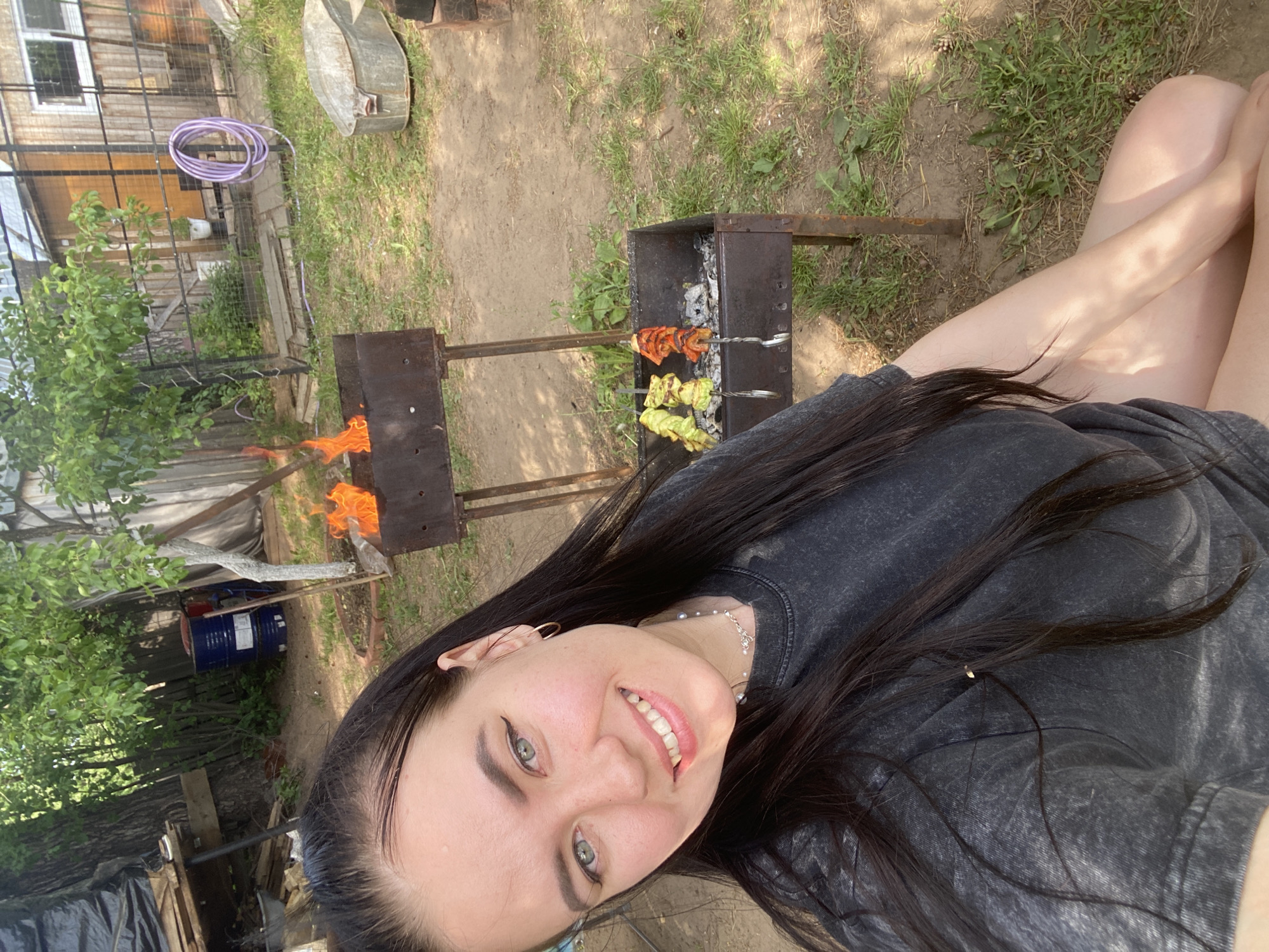 The image shows a woman with dark hair and light eyes, smiling and looking at the camera. She is wearing a dark gray t-shirt and a necklace. Behind her is a backyard setting. There is a grill with flames and skewers of food. The location appears to be Chaikovsky, Russia, based on details in the surrounding environment. There are trees, a fence, a coiled purple hose, and a galvanized metal container in the background. Other objects in the background include blue barrels, stacked lumber, and a structure covered in dark material.