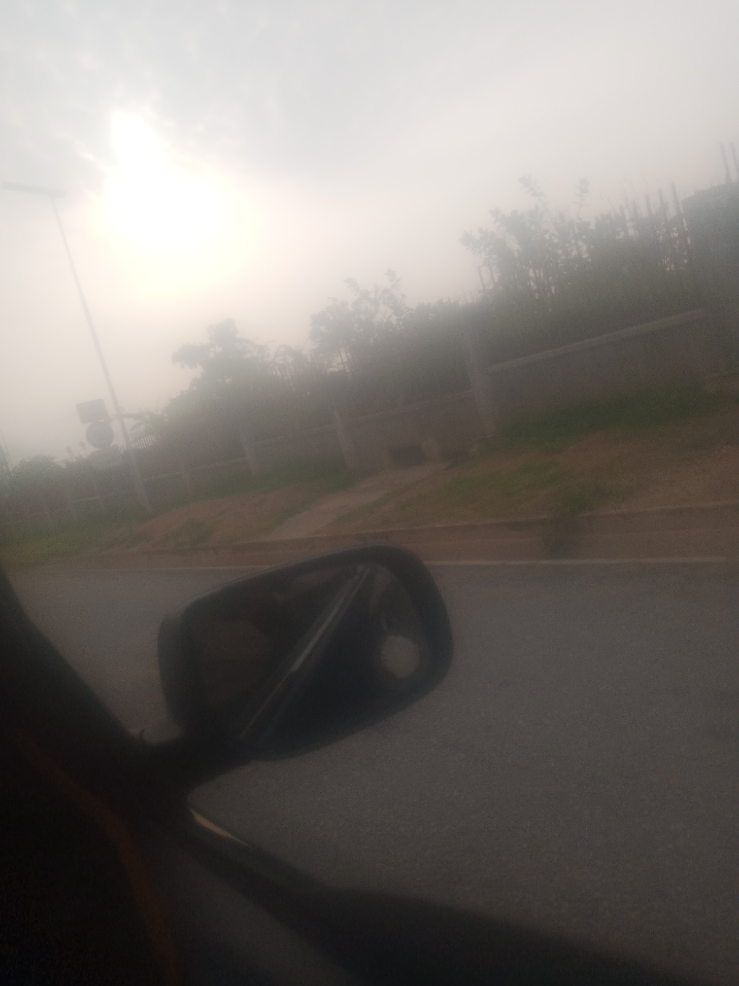 The image captures a hazy view from inside a vehicle, looking out towards a street and roadside. The sky is overcast and appears foggy or dusty, with the sun visible as a bright, diffused light source. On the right side of the image, a side-view mirror of the vehicle is prominent in the foreground. Beyond the mirror, a road is visible, with a strip of dry, sandy earth and some sparse green vegetation along its edge. A low, light-colored wall runs parallel to the road, with a line of trees or dense foliage behind it. In the distance, a street light can be seen on the left. The overall impression is one of a dry, possibly dusty or hazy environment, common in certain parts of Jalingo, Nigeria, particularly during the dry season. There are no people clearly visible in the scene.