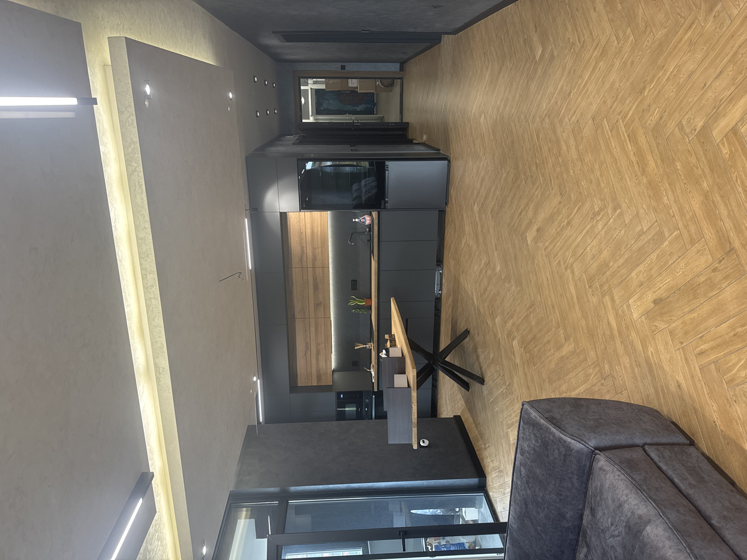 This is an interior photograph of a modern, open-plan living space, likely in Lukovit, Bulgaria, showcasing a combined kitchen, dining, and living area. The image is rotated, displaying the room vertically.
No people are present in the scene. The primary subjects are the architectural and design elements of the room. A sleek, dark gray kitchen occupies the mid-ground, featuring contemporary cabinets, integrated appliances (including an oven and refrigerator), and a wooden backsplash. A kitchen island or bar, with a light wooden top and distinctive dark, criss-cross metal legs, extends into the room. Small decorative items, possibly fruit or a vase, are visible on the kitchen counter.
In the foreground, the back and armrest of a plush, dark gray upholstered sofa are visible, indicating a seating area. The flooring throughout the space consists of light-colored wooden planks laid in a classic herringbone pattern. The walls and ceiling are painted in light, neutral tones, with some sections featuring textured finishes. Extensive modern lighting is integrated into the design, including recessed spotlights in the ceiling, elongated strip lighting along architectural ledges, and under-cabinet lighting in the kitchen, providing a bright and inviting ambiance.
A dark doorway or hallway is visible in the background, leading to other parts of the residence. A reflection or artwork can be partially seen on a wall beyond the kitchen. A large window or glass door is discernible in the bottom left section of the image (when oriented correctly), suggesting access to natural light, though the interior lighting is prominent.
The scene depicts a clean, well-lit, and stylish modern interior. The focus is on the design and functionality of the space, with no specific activity taking place. The time of day is not apparent, as the artificial lighting dominates the illumination. There is no visible text in the image.