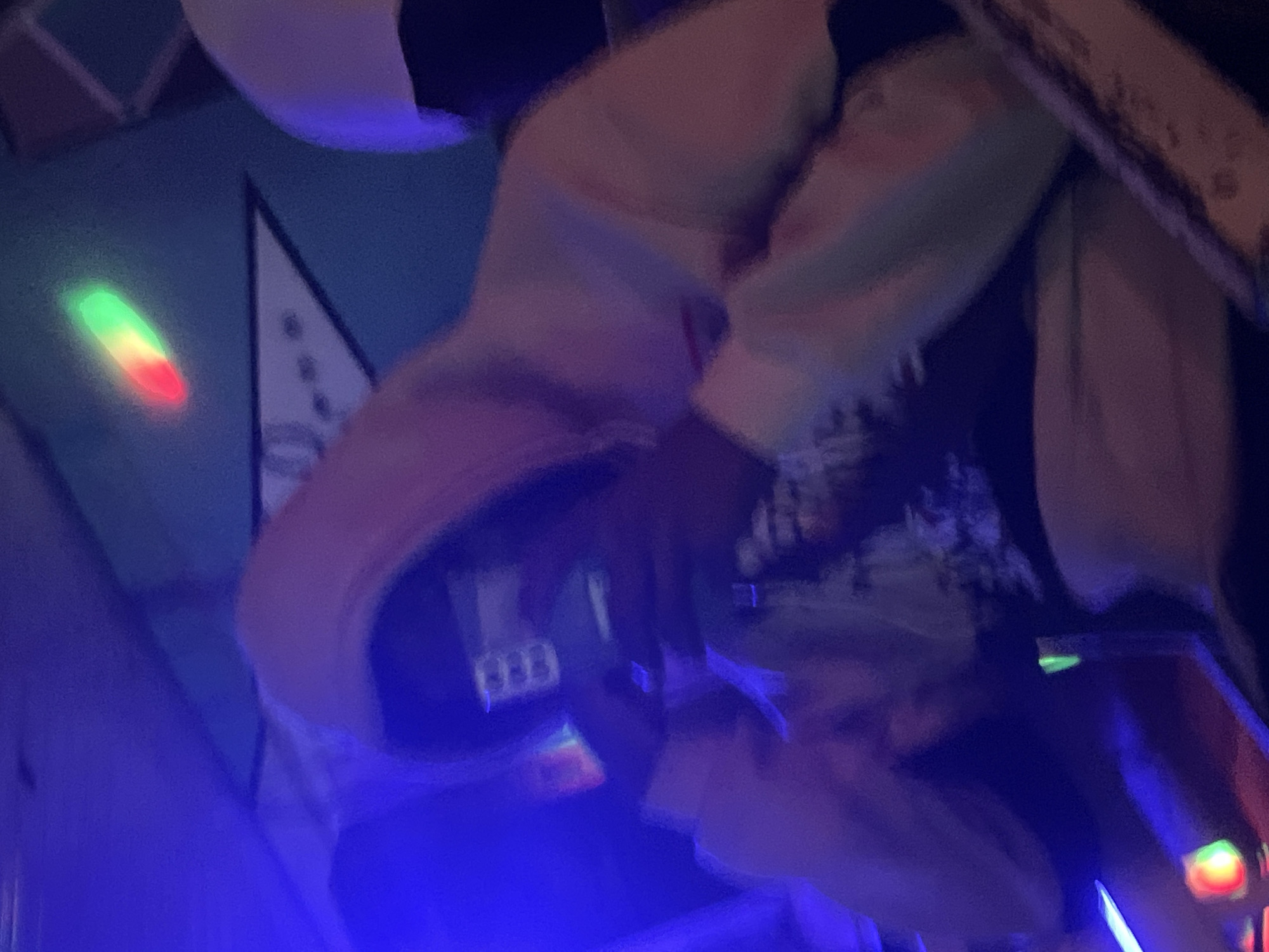 The image shows two individuals in a dimly lit indoor environment, possibly a club or entertainment venue, with an emphasis on blue and purple lighting. The person in the foreground, wearing a light-colored hooded sweatshirt, is holding a smartphone and appears to be taking a picture or video. The person's face is obscured by the phone and the hood of the sweatshirt. Another individual, also wearing a hooded garment, is visible in the background. A triangular graphic with circular elements and a red, green, and yellow light streak are visible on the wall in the background. The scene is characterized by low light conditions and the presence of artificial lighting, creating a somewhat abstract and moody atmosphere. The location in Wumat, Nigeria, is not explicitly discernible from the visual information presented in the image.