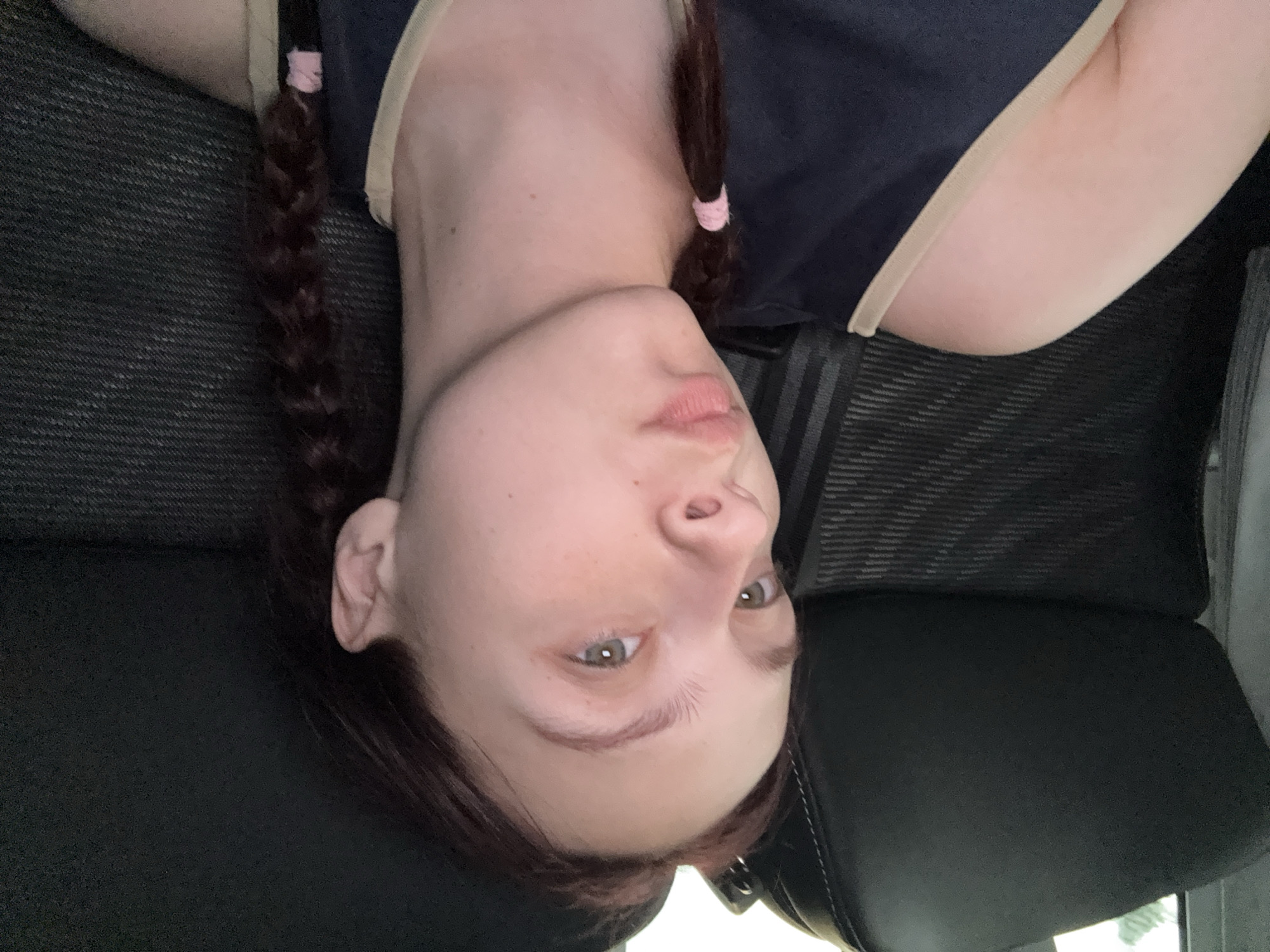 A young adult female with fair skin and reddish-brown hair is positioned in the image. She has two braids held with pink bands. She is wearing a navy blue top with a tan-colored trim. The background suggests an indoor setting, possibly a vehicle, with dark grey patterned fabric and a black leather seat. There is a seatbelt visible. There is no indication of location.