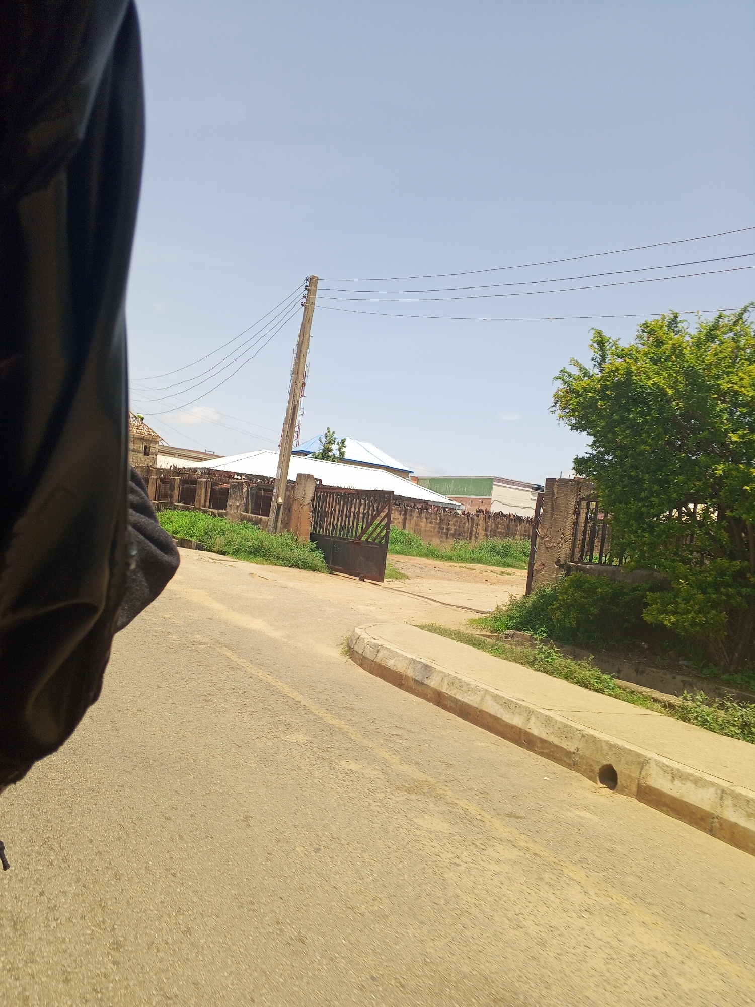 The image depicts a street scene in Kabong, Nigeria. In the foreground, a portion of a dark object, possibly clothing or a vehicle interior, occupies the left side of the frame. The road, paved with asphalt, curves to the right, with a concrete curb and drainage channel visible along its edge.
In the background, several structures are visible. A weathered wooden utility pole with multiple electrical wires stretches across the sky. Adjacent to the pole, a metal gate, partially obscured by vegetation, stands open, leading into a compound. Beyond the gate, a low brick wall with a metal fence tops it forms a boundary. A white-roofed building, potentially a house or other structure, is situated behind the wall, with additional buildings and construction visible further back. Green vegetation, including trees and bushes, is scattered throughout the scene, particularly on the right side and along the perimeter of the compound. The sky is clear and blue, indicating a sunny day. The overall impression is one of a residential or mixed-use area in an urban or semi-urban setting.
