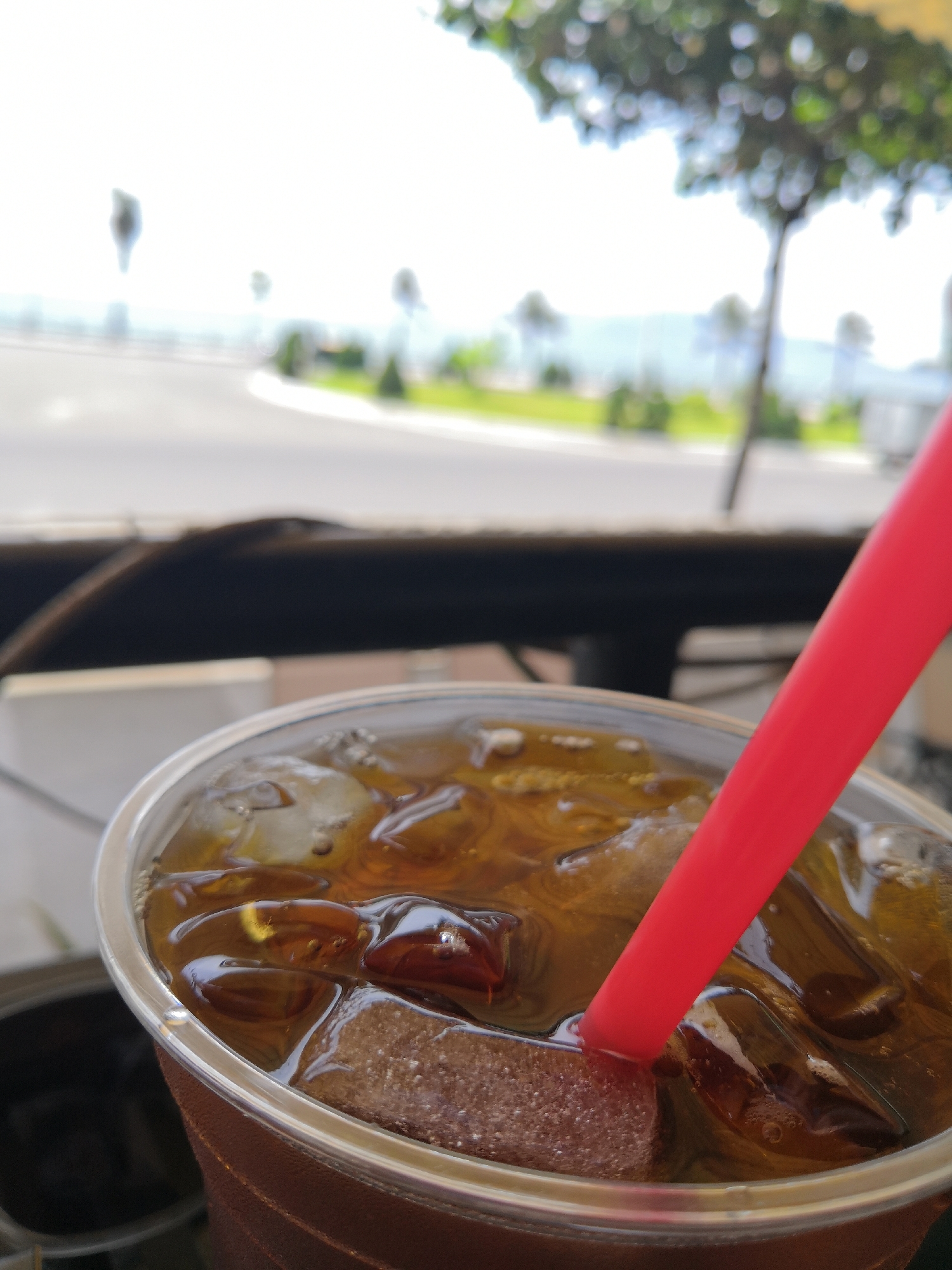 A close-up shot captures a clear plastic cup filled with a dark beverage and ice cubes, a bright red straw submerged within. The cup has a brown outer sleeve. In the background, the scene is softly blurred, suggesting an outdoor environment. A road curves into the distance, bordered by green grass and foliage. In the far background, a hazy view of hills or mountains can be seen under a bright sky. The lighting indicates it is daytime. The location is identified as Bac Nha Trang Ward, Vietnam.