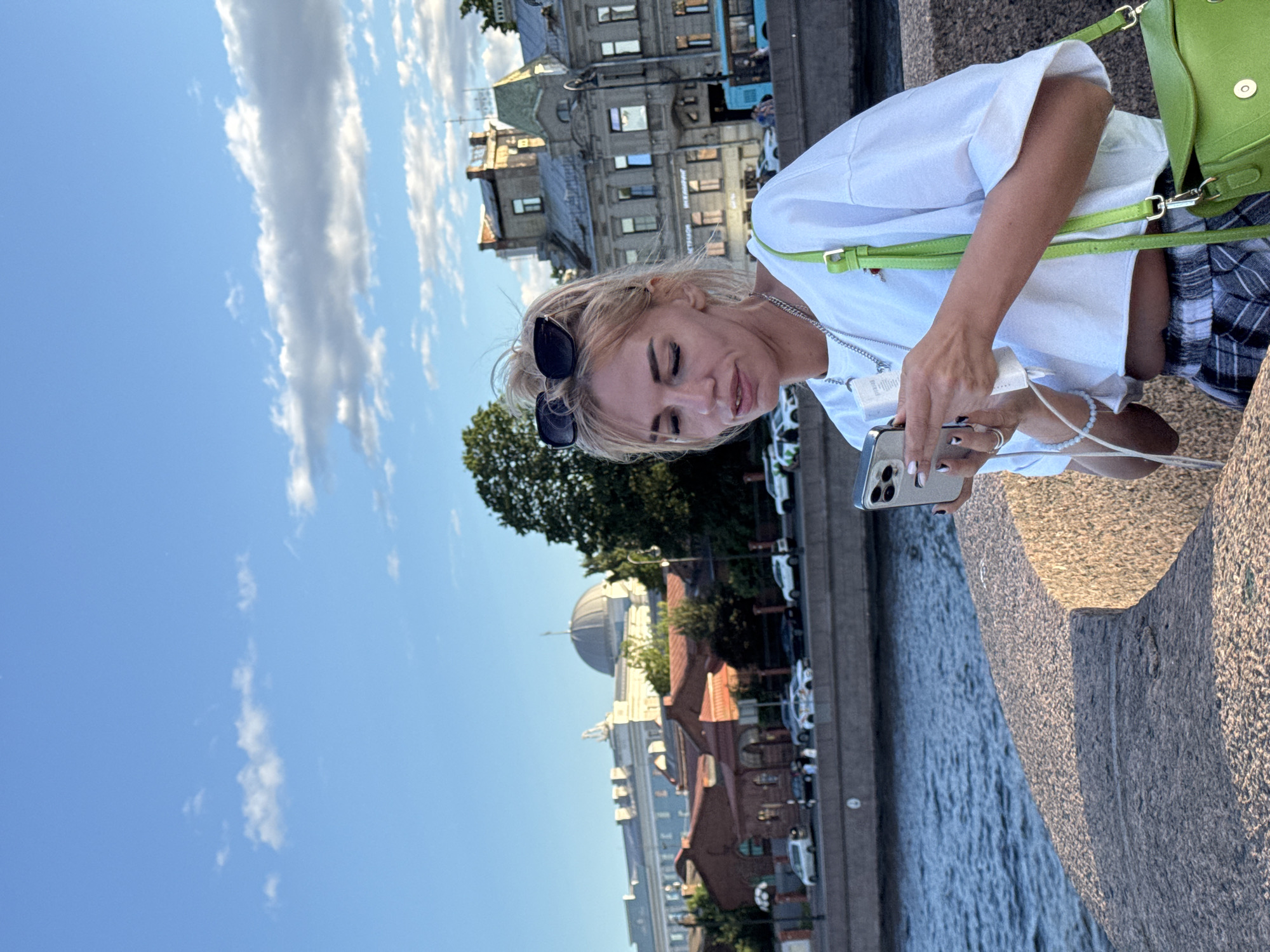 A blonde woman with sunglasses on her head stands on a stone embankment overlooking a body of water, with the city of Saint Petersburg in the background. She is wearing a white crop top, plaid shorts, and a bright green shoulder bag. In her hands, she holds a smartphone and a small white object, appearing to be engrossed in her phone. The background shows historic buildings and a road with parked cars. The sky is blue with scattered clouds, indicating daytime. The scene is set in Saint Petersburg, Russia, likely along one of its many canals or the Neva River.