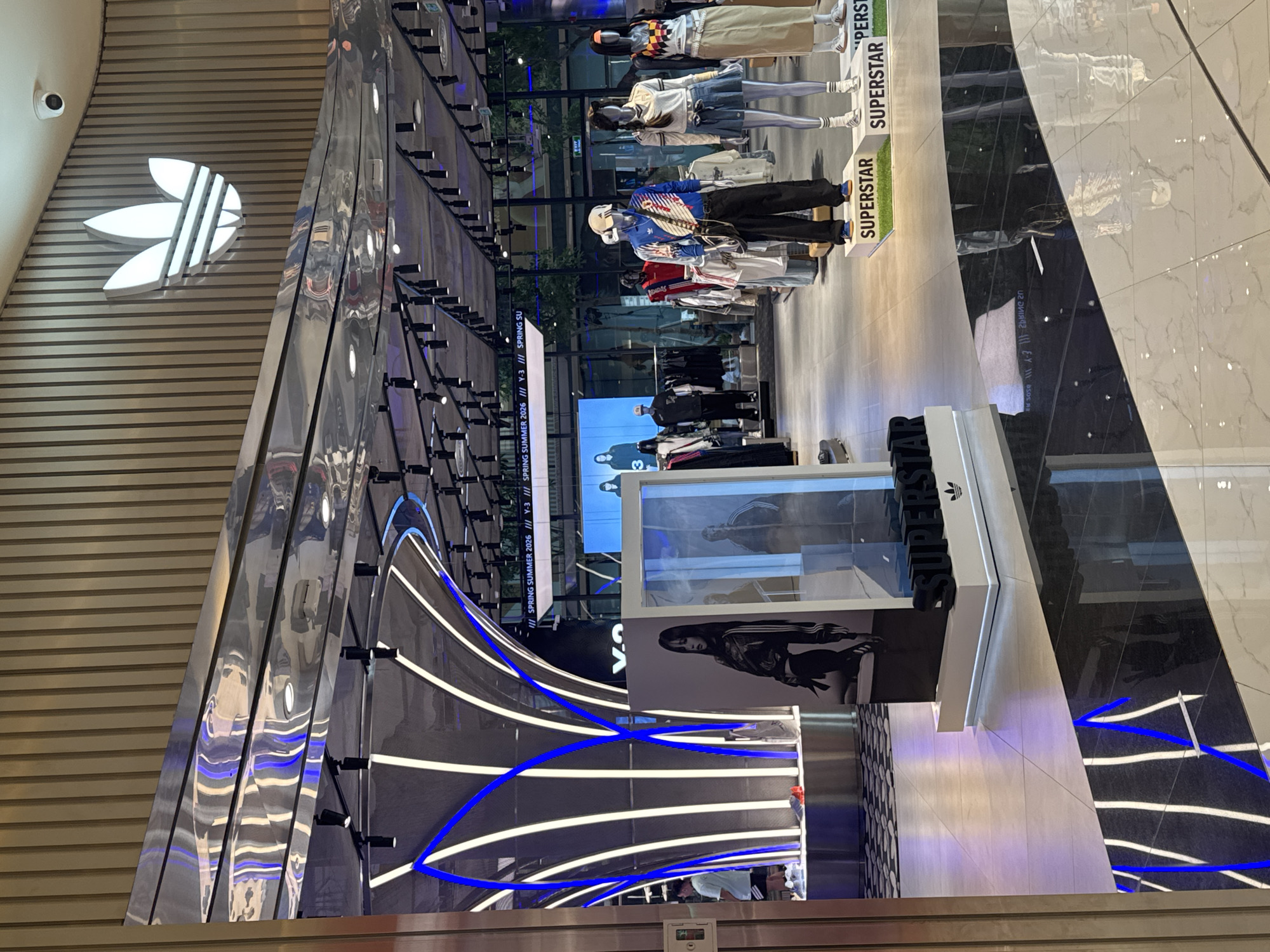 The image depicts the interior of an Adidas store, likely located in a high-end shopping mall in Hà Nội, Vietnam. The store features modern design elements, including a metallic wall with an Adidas logo and an array of clothing displays with mannequins dressed in Adidas apparel. The "SUPERSTAR" model is heavily featured. The flooring is a dark marble-like material, adding to the store's luxurious ambiance. Display stands with additional clothing items are also present. Illumination comes from bright, linear lighting fixtures and spotlights directed at the merchandise.