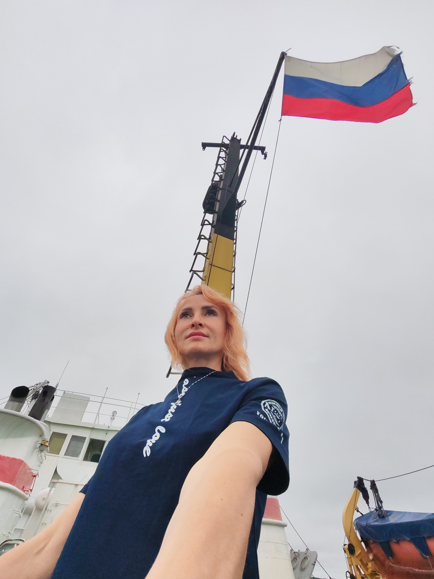A woman with reddish-blonde hair stands on the deck of a ship, looking up at a Russian flag flying from a mast. The sky is overcast. The woman is wearing a dark blue t-shirt with white writing and a silver necklace. The ship's structure is visible behind her, as is a life raft. The location is identified as the city of Novokemskii, Russia.