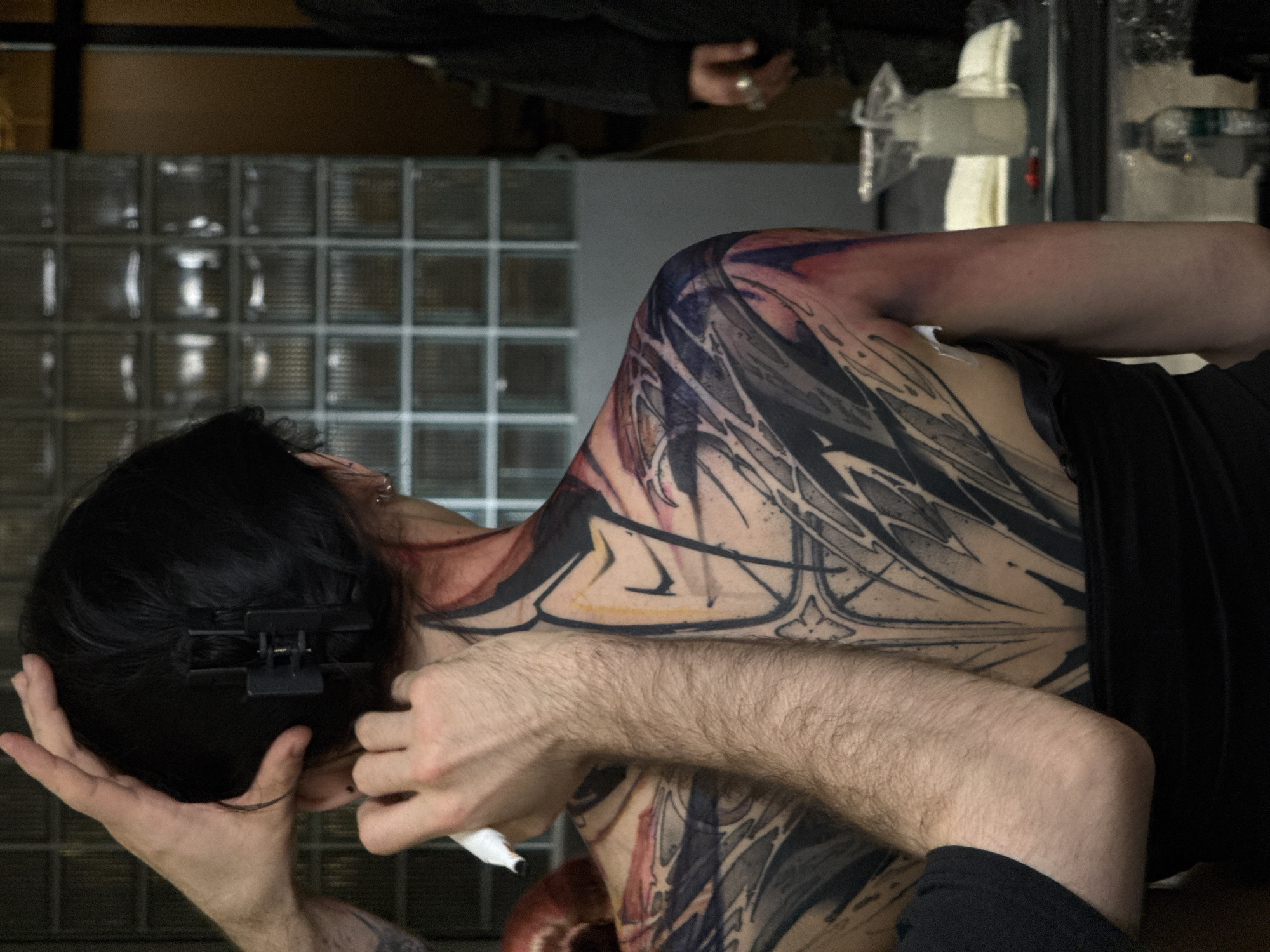 A man is lying on his side, getting a tattoo on his back and shoulder. The tattoo is large and covers most of his back, featuring abstract black, gray, red, and yellow designs. The man has dark hair, pulled back with a black clip, and he is holding a white pen or marker in his hand. His arm and hand are hairy. He is wearing a black top. Behind him is a wall made of glass blocks. There are other people and tattoo equipment visible in the background. The location appears to be a tattoo studio, not specifically identifiable as Moscow.