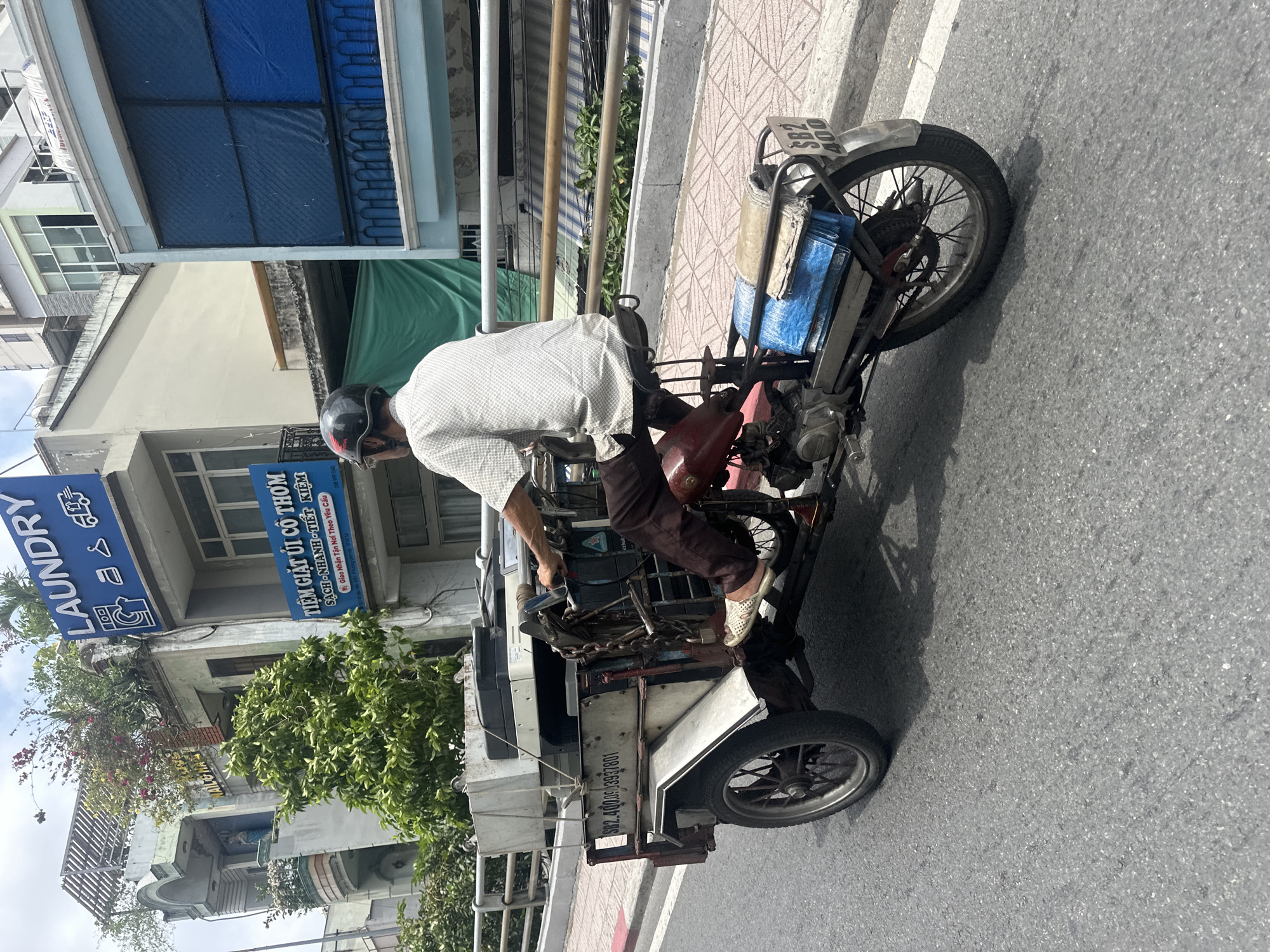 An adult male, wearing a dark helmet with a Honda logo, a light patterned short-sleeve shirt, dark trousers, and light woven slip-on shoes, operates a customized three-wheeled motorized cargo vehicle. The driver is seated on a saddle, facing away from the camera, traversing an asphalt road.
The vehicle consists of a motorcycle front section with a red fuel tank and visible engine components, modified to incorporate a white, two-wheeled cargo bed. The cargo bed transports a large white office copier/printer, secured by ropes. A heavy chain with a padlock is visibly attached to the vehicle's frame near the driver. Inscriptions on the side of the cargo bed include "SB2.400.009397801." The motorcycle's rear fender displays a partial license plate, "59-S2 400." A blue tarp and other rolled materials are strapped to the motorcycle's rear rack.
The immediate environment features a patterned red-tiled sidewalk and a silver-colored metal railing running parallel to the road. The background reveals urban architecture in Thủ Đức, Vietnam, under an overcast sky. A prominent blue sign on a multi-story building displays "LAUNDRY" in white text with washing machine, iron, and delivery truck icons. Below it, a smaller blue sign reads "TIỆM GIẶT ỦI CÔ THƠM" (Cô Thơm Laundry Shop), indicating services as "Sạch - Nhanh - Tiết Kiệm" (Clean - Fast - Economical) and "Giao Nhận Tận Nơi Theo Yêu Cầu" (Delivery at your request). This laundry business is located in Thủ Đức, Thành phố Hồ Chí Minh, Vietnam, with a known address such as 122 Đ. Số 8, Linh Xuân. Adjacent buildings feature various facades, balconies, and extensive rooftop and balcony vegetation, including flowering plants.