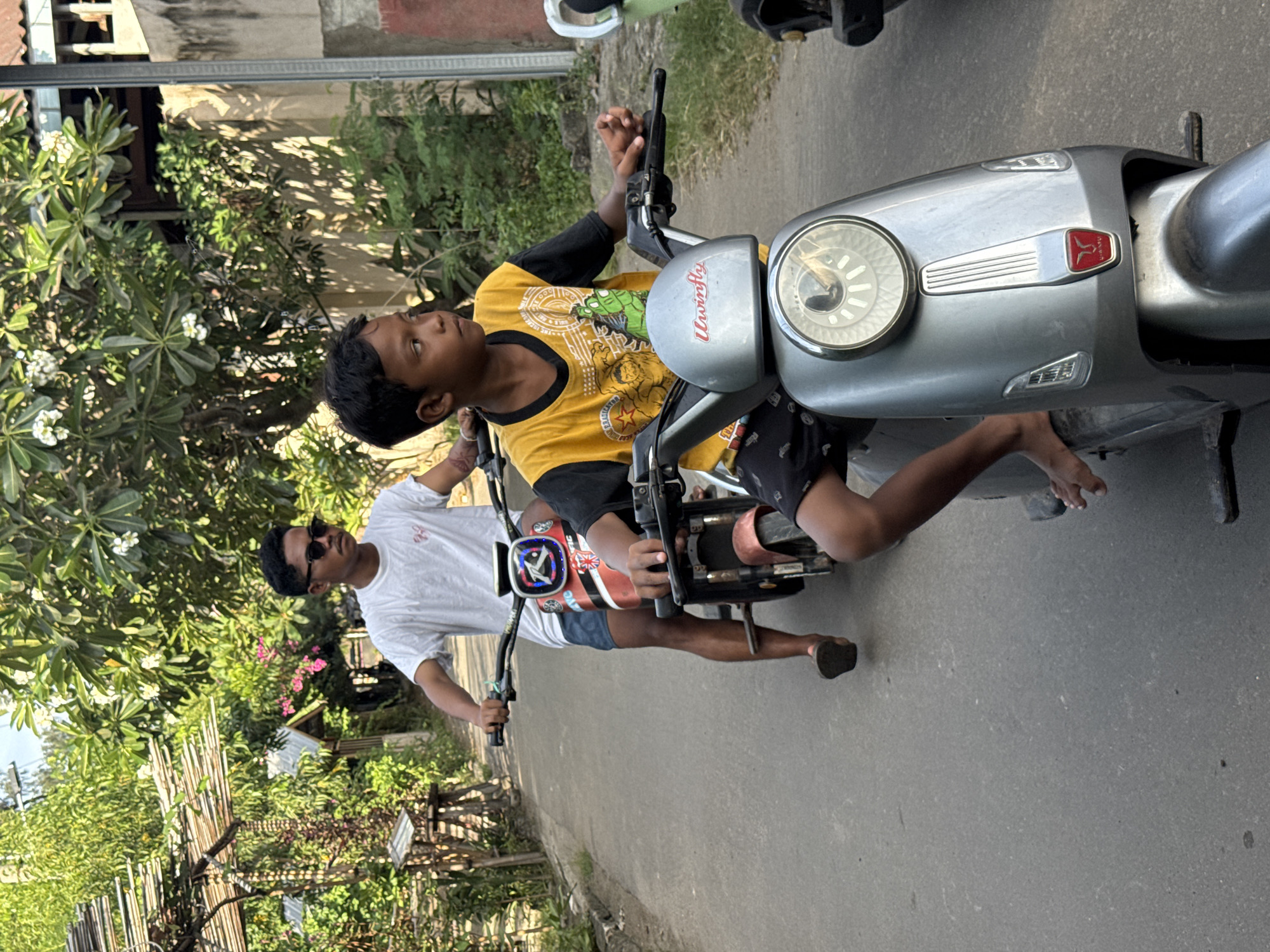 The image shows an adult male with dark hair and sunglasses, wearing a white t-shirt and blue shorts, driving a scooter with a child on the front. The child, wearing a yellow shirt with black sleeves and shorts, is holding onto the handlebars. They are on a paved road, with lush greenery visible on both sides, including trees and flowering plants. Buildings and fences are also visible in the background. The scooter is gray with a red logo. The location is Unknown, Indonesia, based on the metadata.