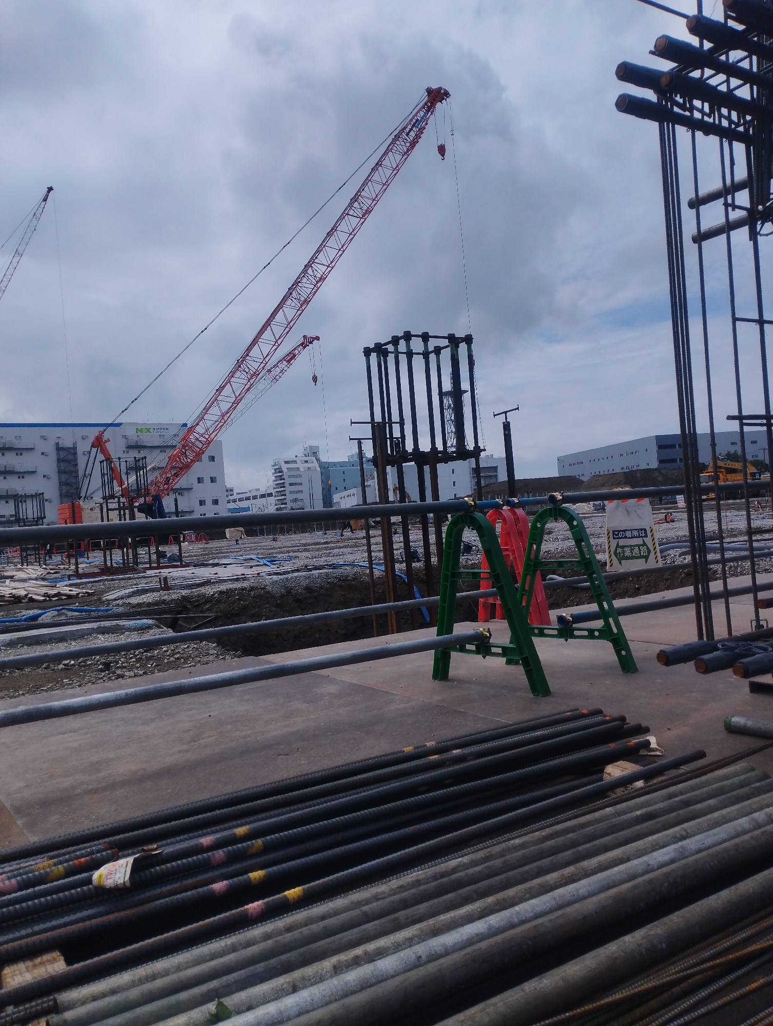 This is a construction site in Ichikawa, Japan, with a crane in the background and rebar in the foreground. The site is covered in dirt and gravel, with some temporary structures and equipment visible. The sky is cloudy and overcast. There are no people visible in the image.
