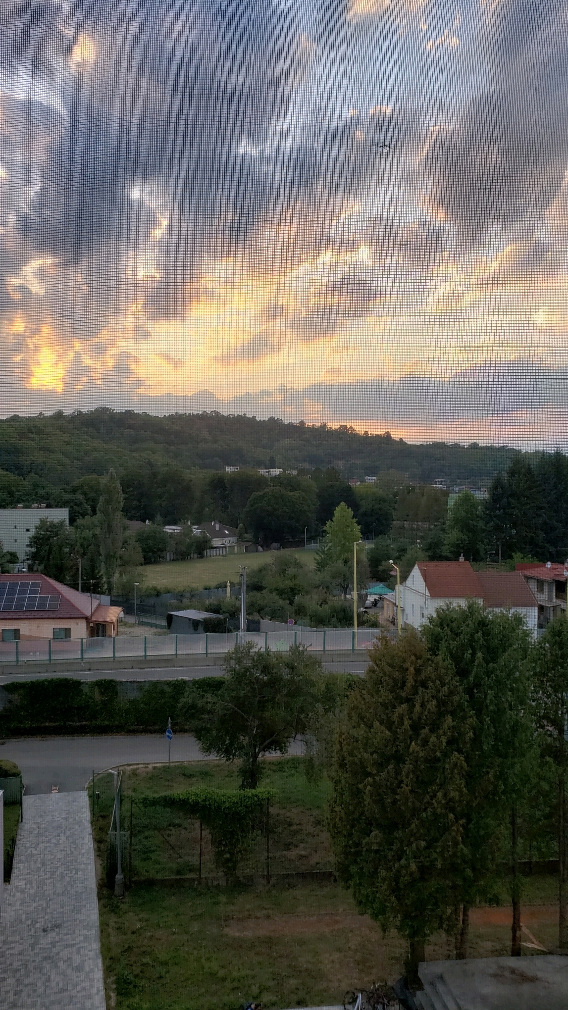 This is a vertical, eye-level, outdoor, daytime photograph taken through a window screen, showing a view of a suburban or semi-rural area at sunset. The sky is filled with dramatic clouds in shades of gray, blue, and orange, with the sun's golden light breaking through the clouds on the left side of the frame. Below the sky is a rolling landscape of green hills covered in trees, with scattered buildings and houses visible among the foliage. In the foreground, there is a street or road with a low, transparent barrier on the right. To the left of the road, there is a building with a red tiled roof and solar panels. Further left, a pedestrian walkway made of light-colored paving stones leads into the scene. Several trees are scattered throughout the middle and foreground. The overall impression is a peaceful, natural setting with signs of human habitation. The exact location is unknown.