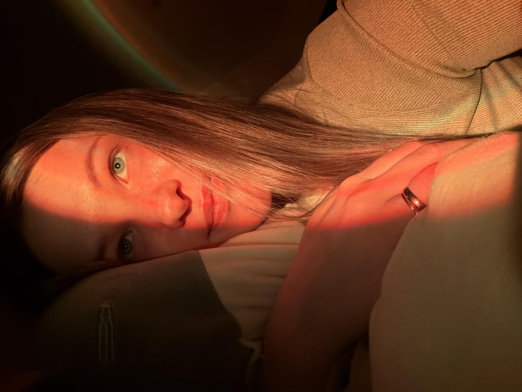 A woman lies on her side in a dimly lit space. Light casts a warm orange-red glow across her face and hand. She has fair skin, light brown hair, and blue eyes. A ring is visible on a finger of her right hand. She is wearing a beige, textured garment. The environment is shadowy and undefined. There is no information regarding the location.
