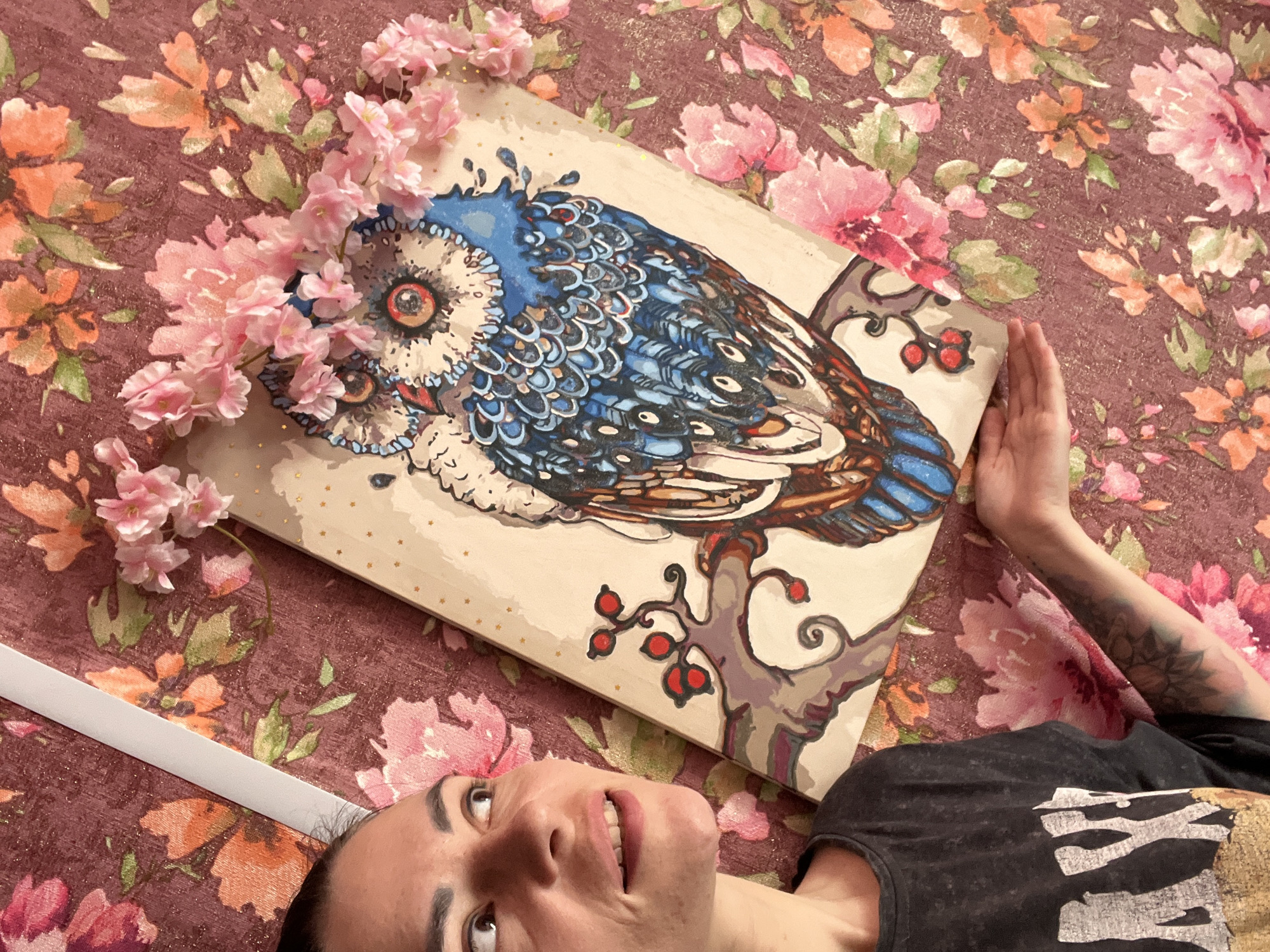 In this overhead shot, a woman lies on her back on a floral-patterned surface, looking up and to her right with a slight smile. A painting of an owl perched on a branch is laid out diagonally on top of her, adorned with artificial pink flowers. The woman is wearing a dark grey t-shirt with a white graphic design. Her arm, visible on the right side of the frame, has tattoos. The background fabric has a deep reddish-purple hue with an impressionistic floral print in shades of pink, coral, and green. The painting itself is rendered on a light-colored wood panel and features a blue and white owl with striking red eyes, surrounded by small golden dots. The overall mood is artistic and relaxed, with the subjects integrated into a visually rich environment. No specific time of day or weather conditions are indicated. The location context of Krasnoe-on-Volga, Russia, is provided.